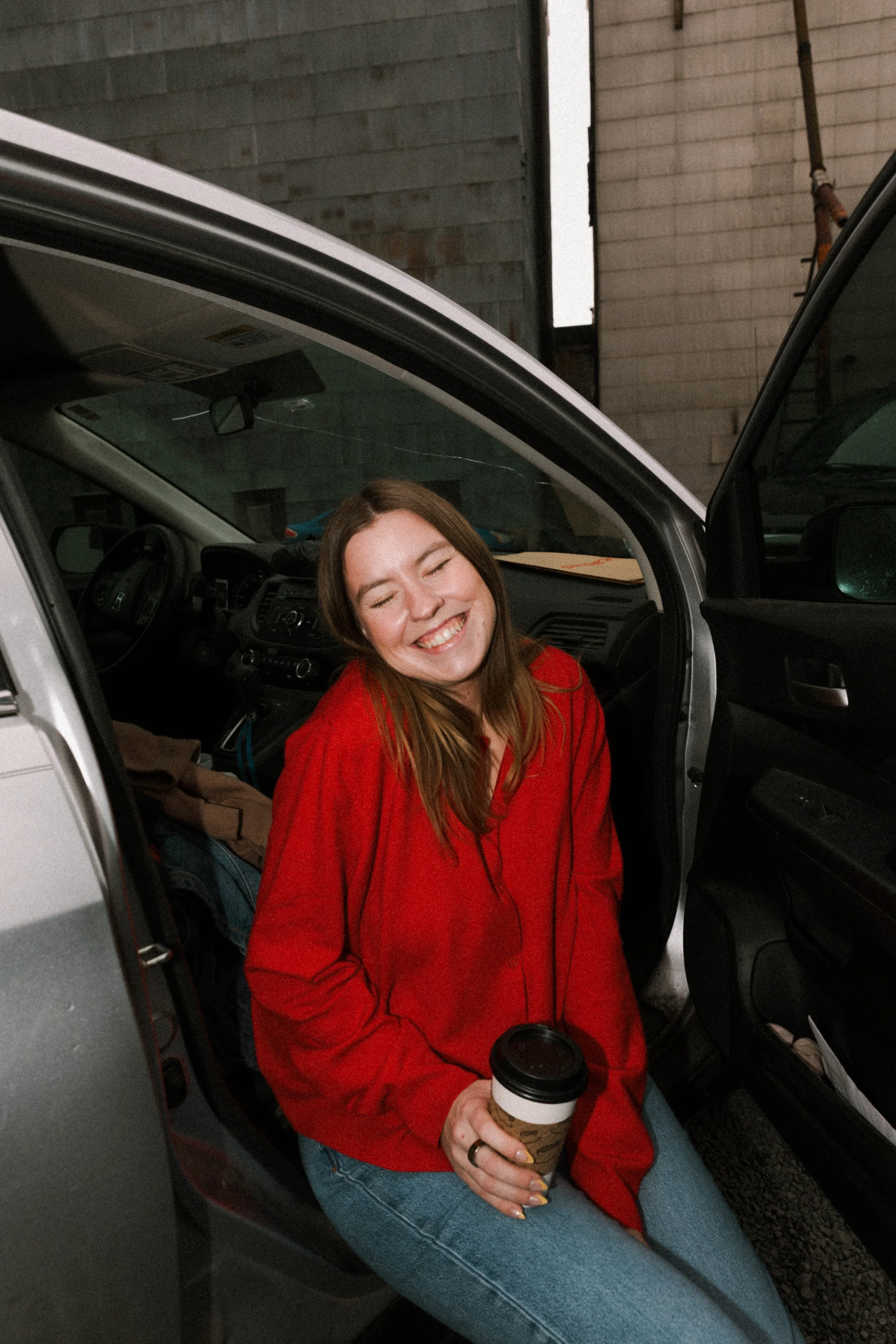 A woman in a red jacket sitting in the passenger seat of a car, smiling with eyes closed, holding a coffee cup, with a background of a brick wall.