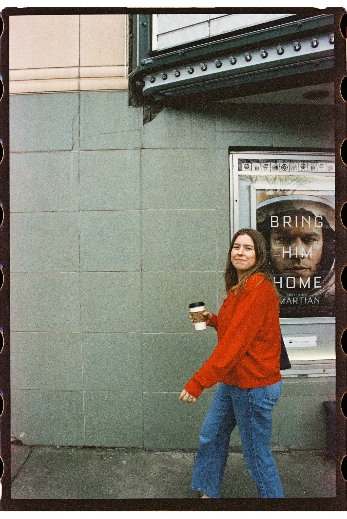 A woman in a red jacket holding a coffee cup walking past a building that has a poster with the text 'Bring Him Home' and an image of a man's face.