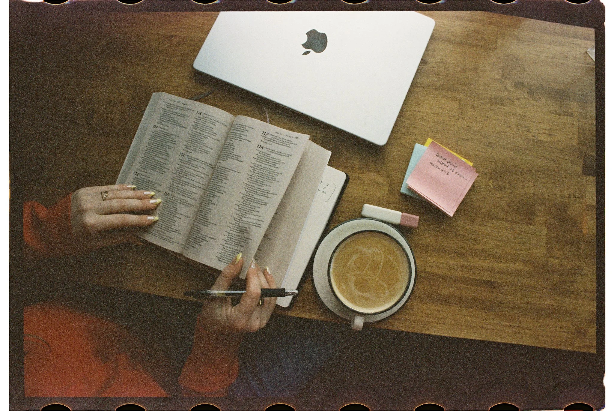 Top-down view of a wooden desk with a closed silver MacBook, an open book, a coffee mug with latte, a hand holding a pen, sticky notes, and a whiteboard marker.
