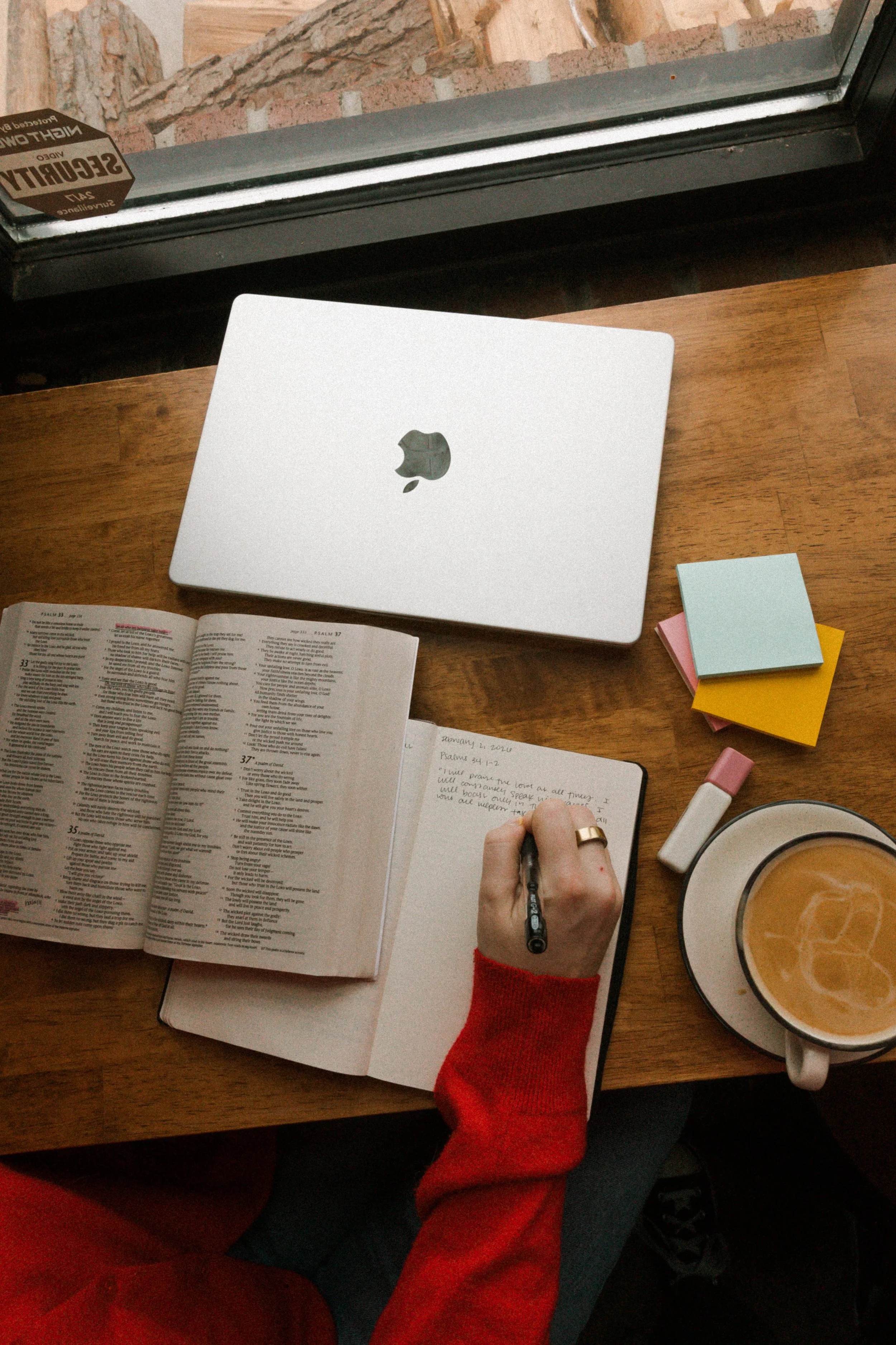 A person wearing a red sweater writing in a notebook at a wooden table next to a closed silver MacBook and a cup of coffee. There are sticky notes, a highlighter, and a bible open on the table, with a window in the background.