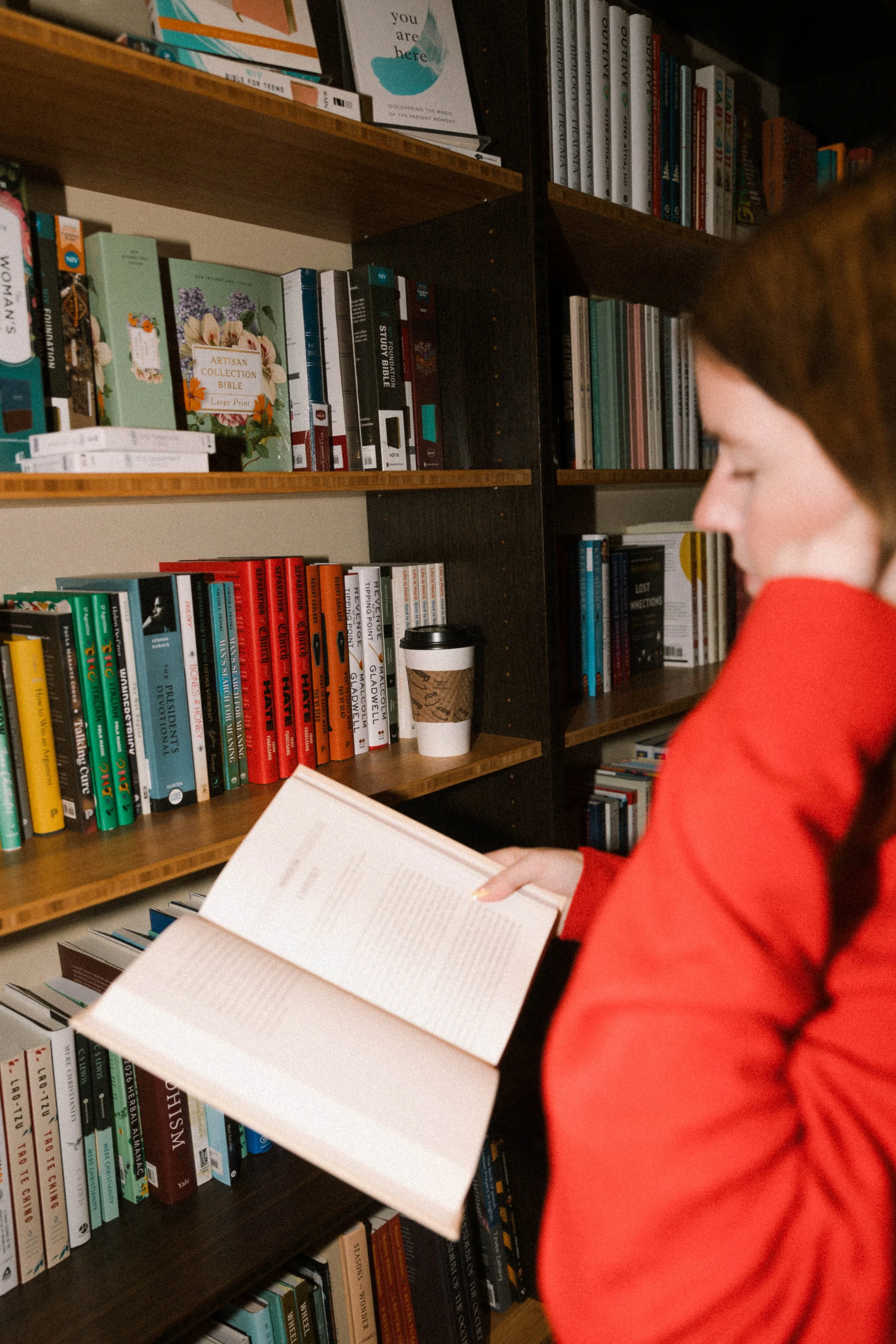 A woman wearing a red jacket is holding and reading an open book in a library aisle with bookshelves filled with various books behind her.