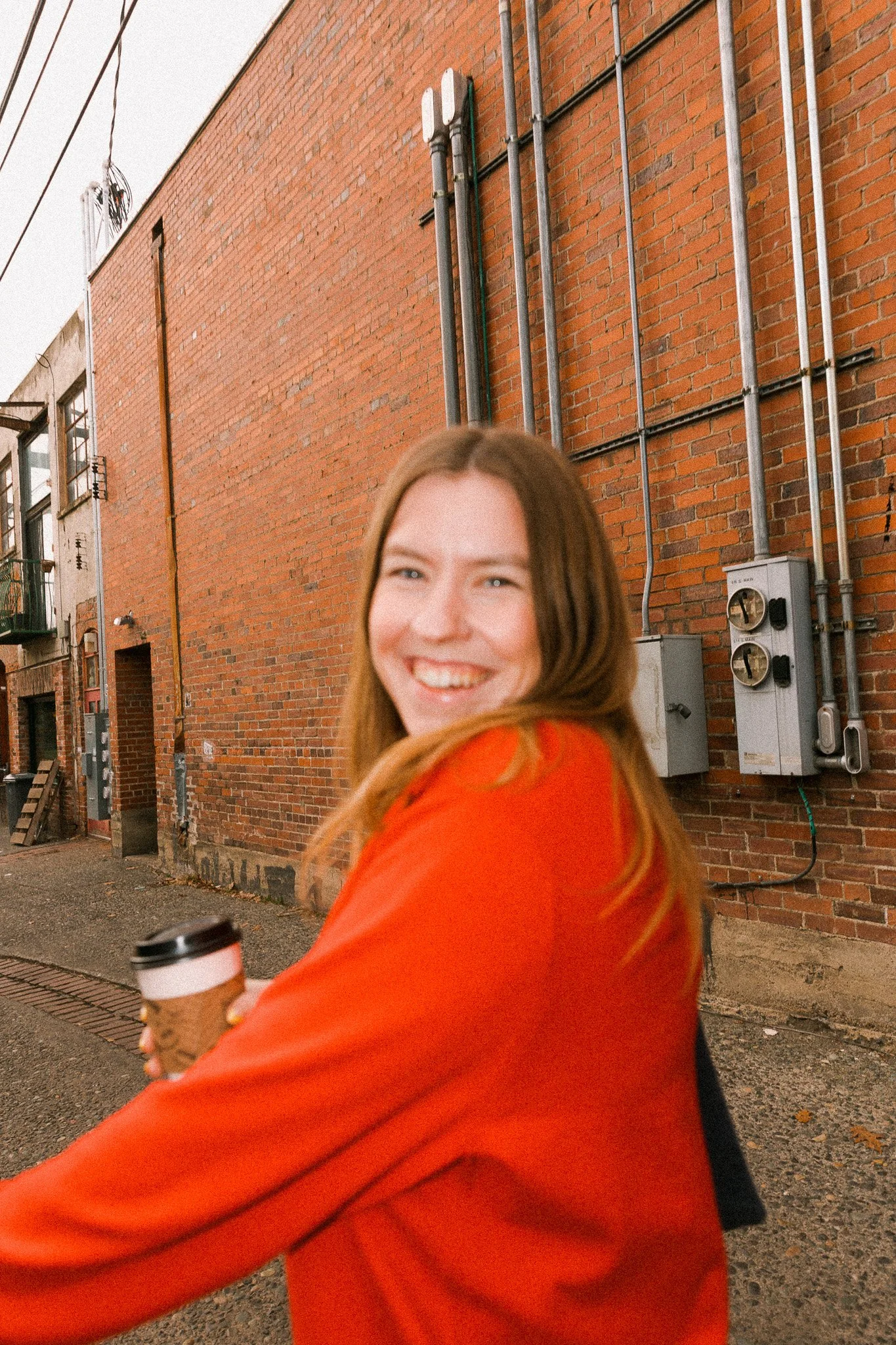 A smiling woman with long reddish hair wearing an orange jacket and holding a disposable coffee cup stands outside against a brick wall with electrical pipes and meters.