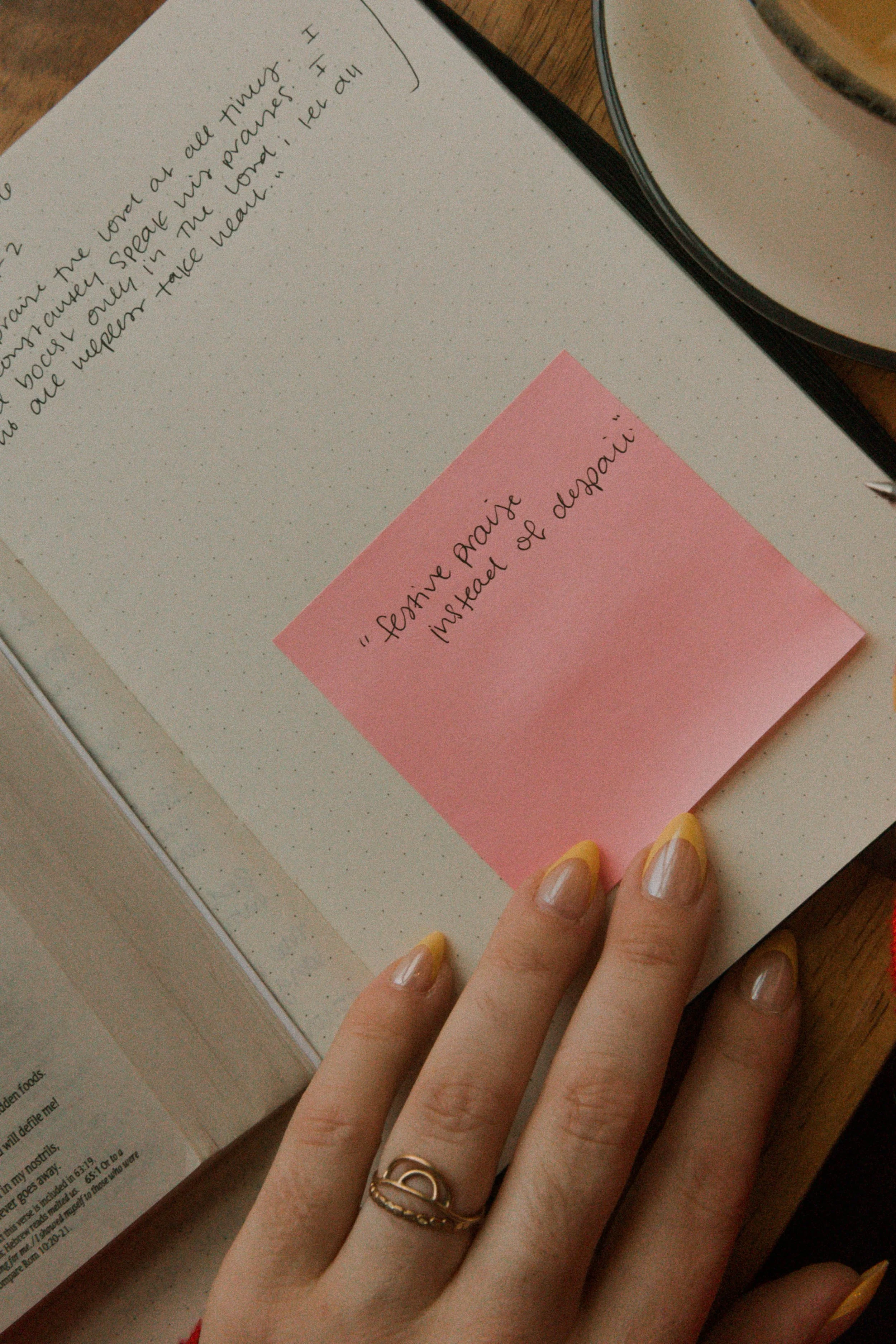 Close-up of a notebook with a pink sticky note that reads "Seemée Prairie installed & despair". A hand with yellow-tipped nails and a gold ring is resting on the notebook.