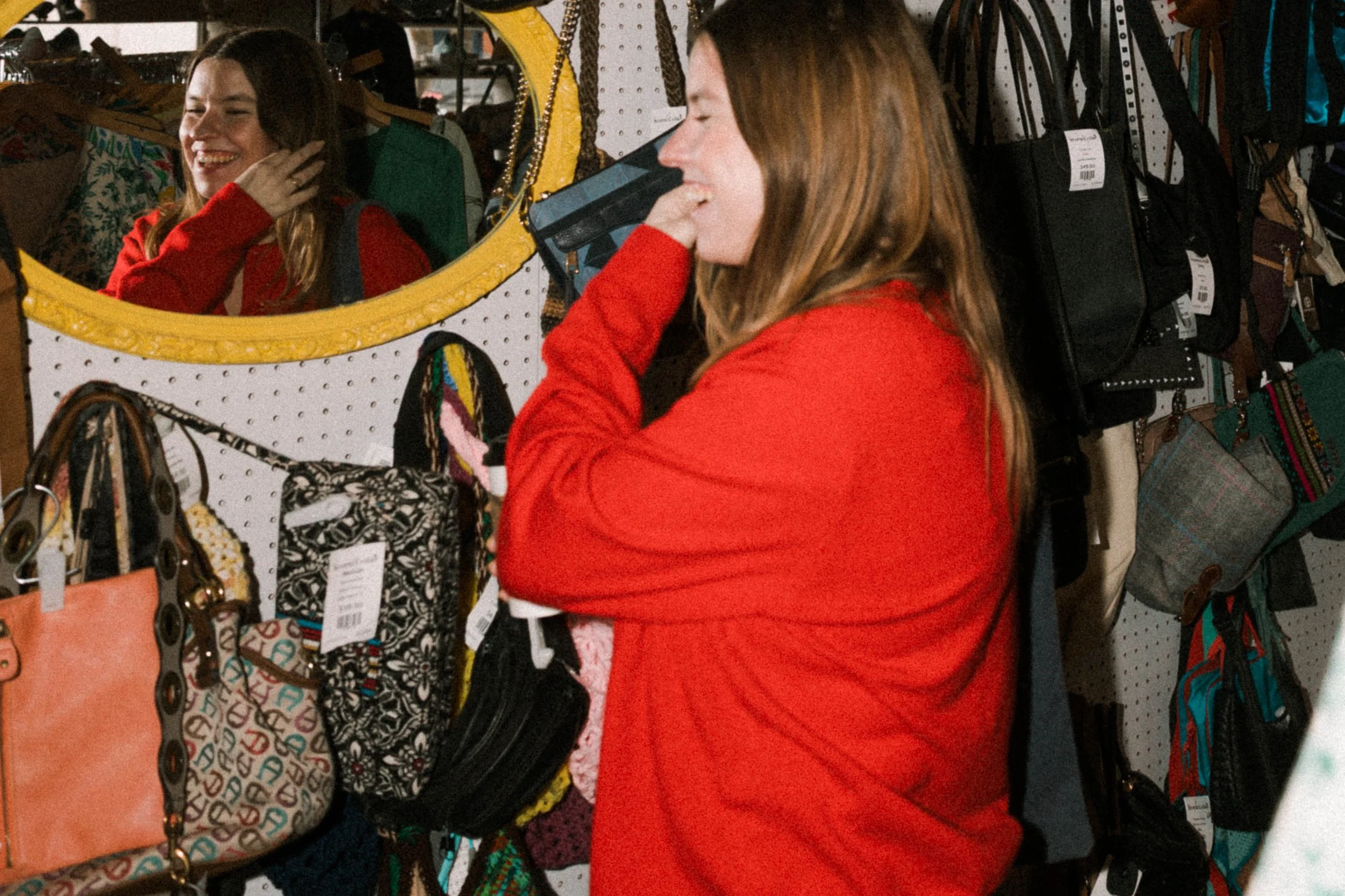 A woman in a red sweater shopping for purses and bags at a store, looking at a bag while smiling and looking into a mirror, with reflection showing her face and smiling