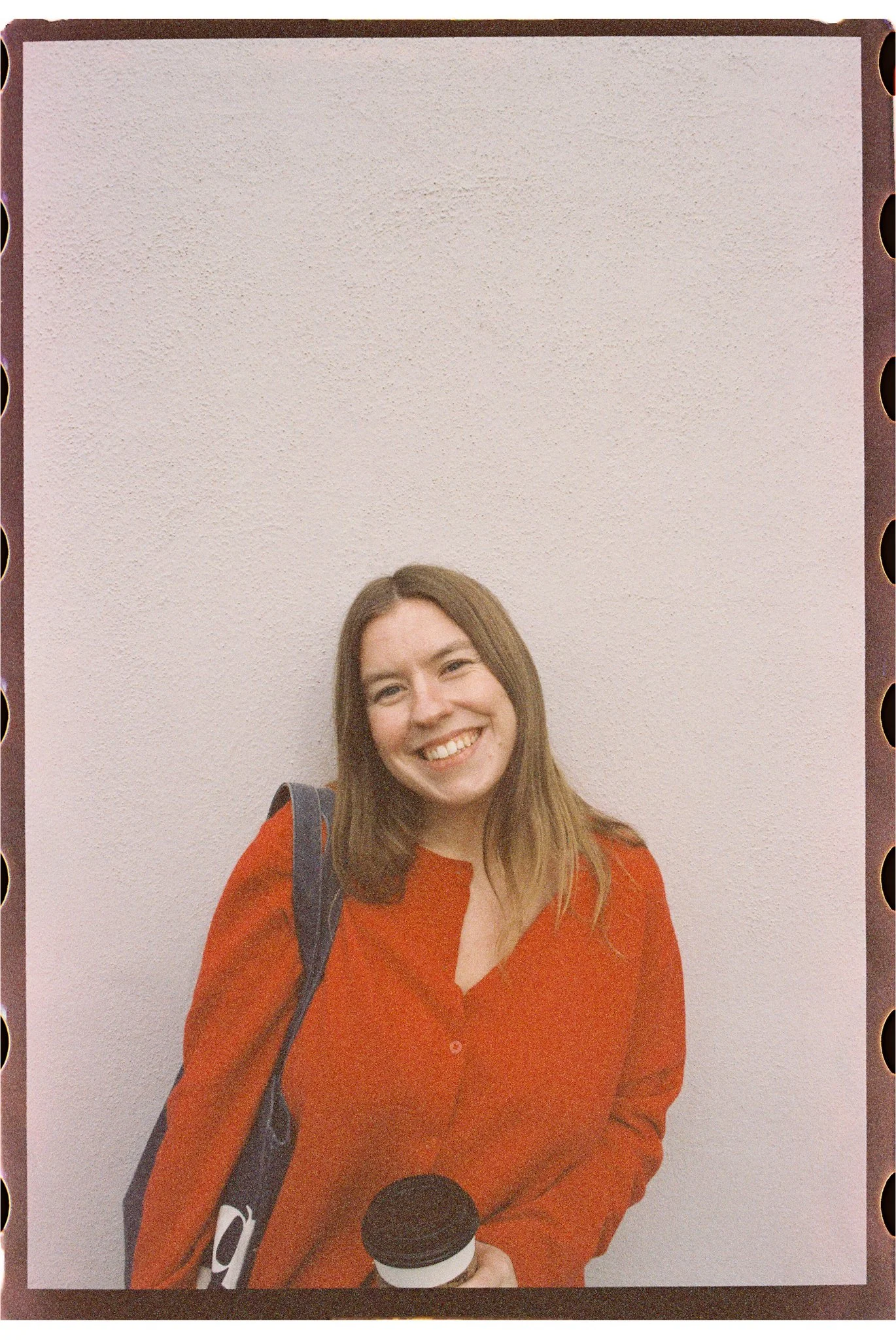 A smiling woman with long brown hair, wearing a red top, holding a coffee cup, standing against a plain white wall.