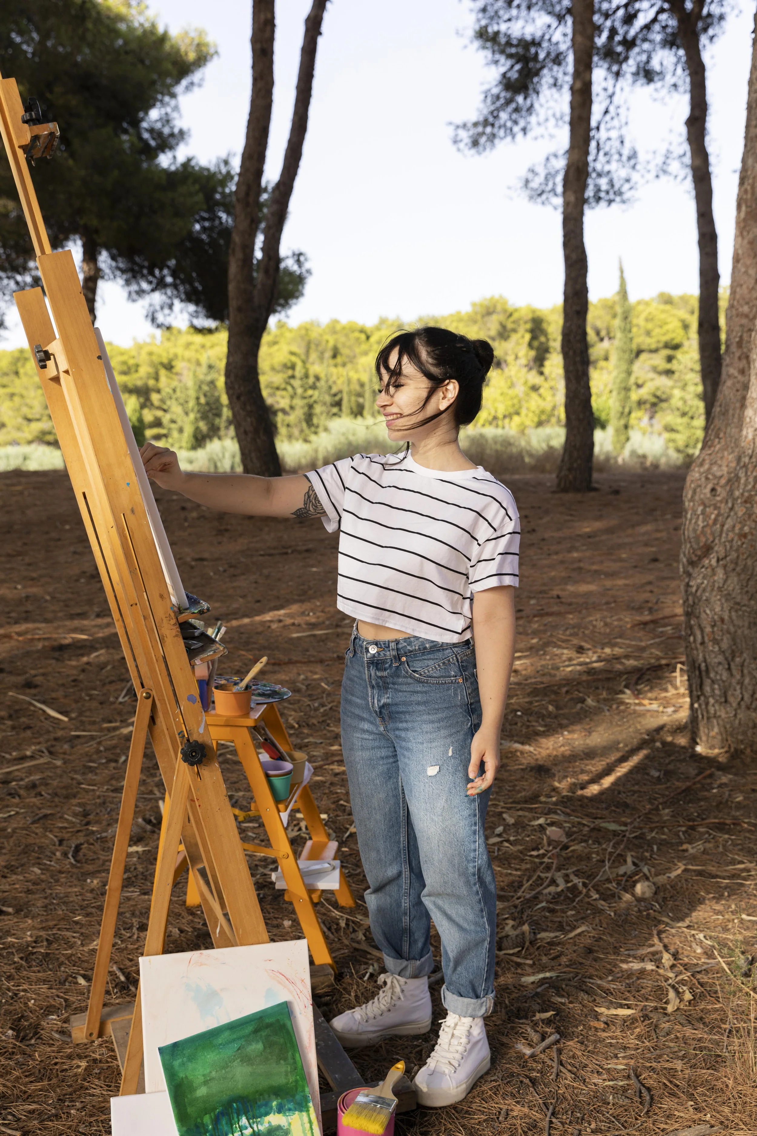 A young woman with dark hair tied in a bun, wearing a striped t-shirt and ripped jeans, is painting outdoors in a wooded area. She is standing in front of an easel with painting supplies, smiling as she works.