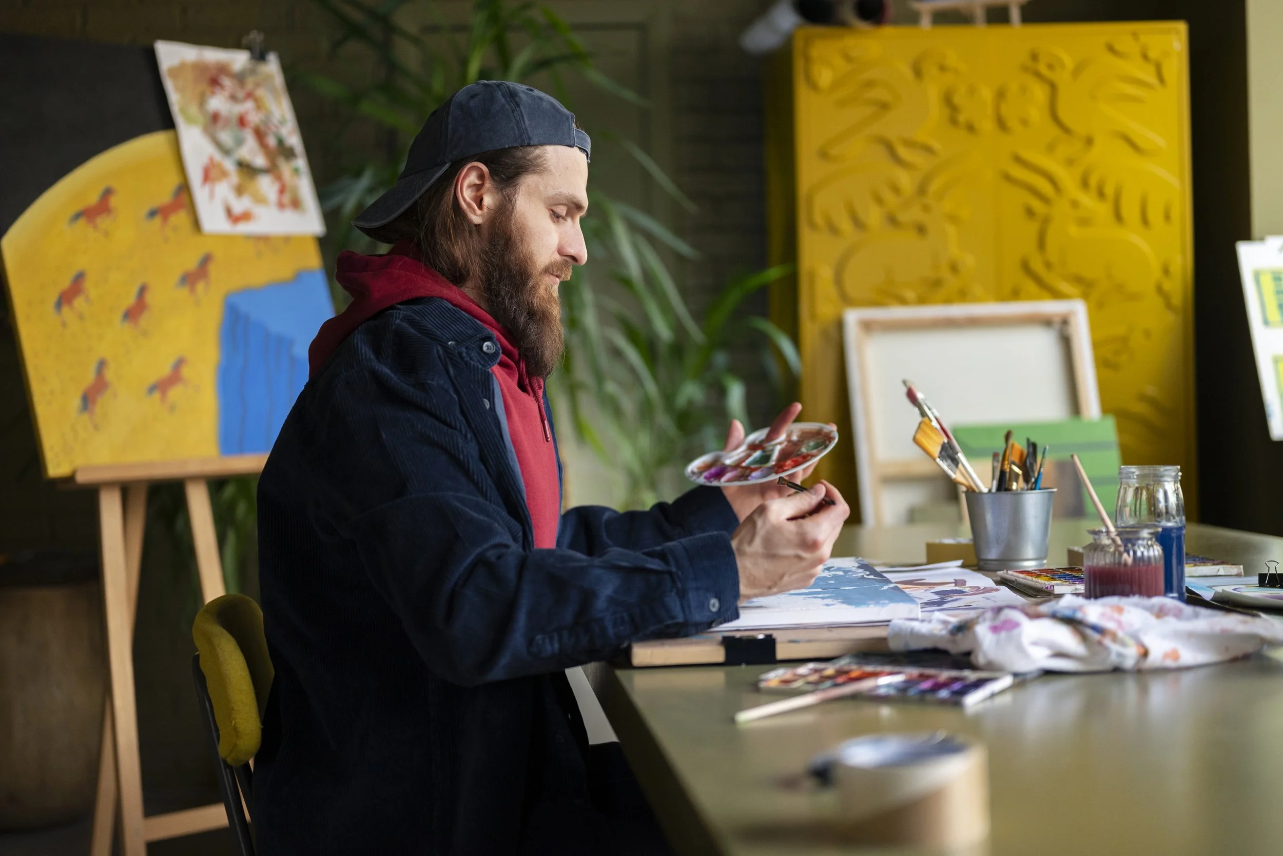 A man with long hair, beard, wearing a backwards cap and a dark jacket, sitting at a table in an art studio, painting with a palette and brushes, with artworks and supplies around him.