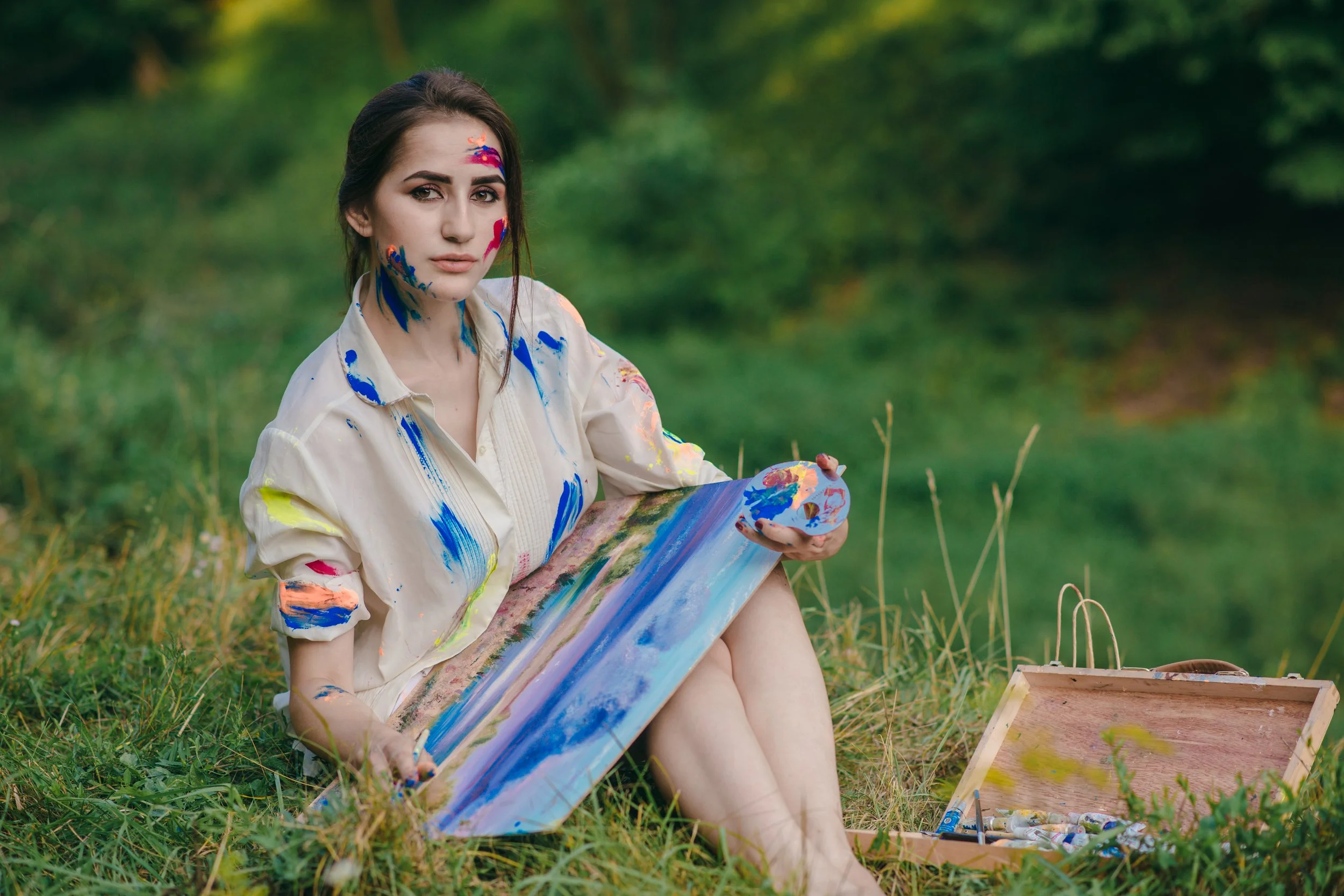 Young woman sitting outdoors on grass holds a painting palette and canvas with a landscape scene. She wears a white shirt splattered with bright paint, and has paint on her face and neck. An open wooden box of paints is beside her.