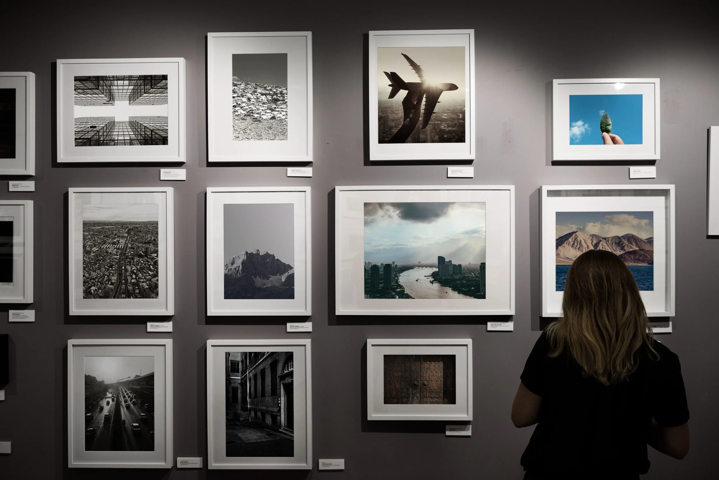 A woman with brown hair looking at framed photographs on an art gallery wall, with some images in black and white and others in color.