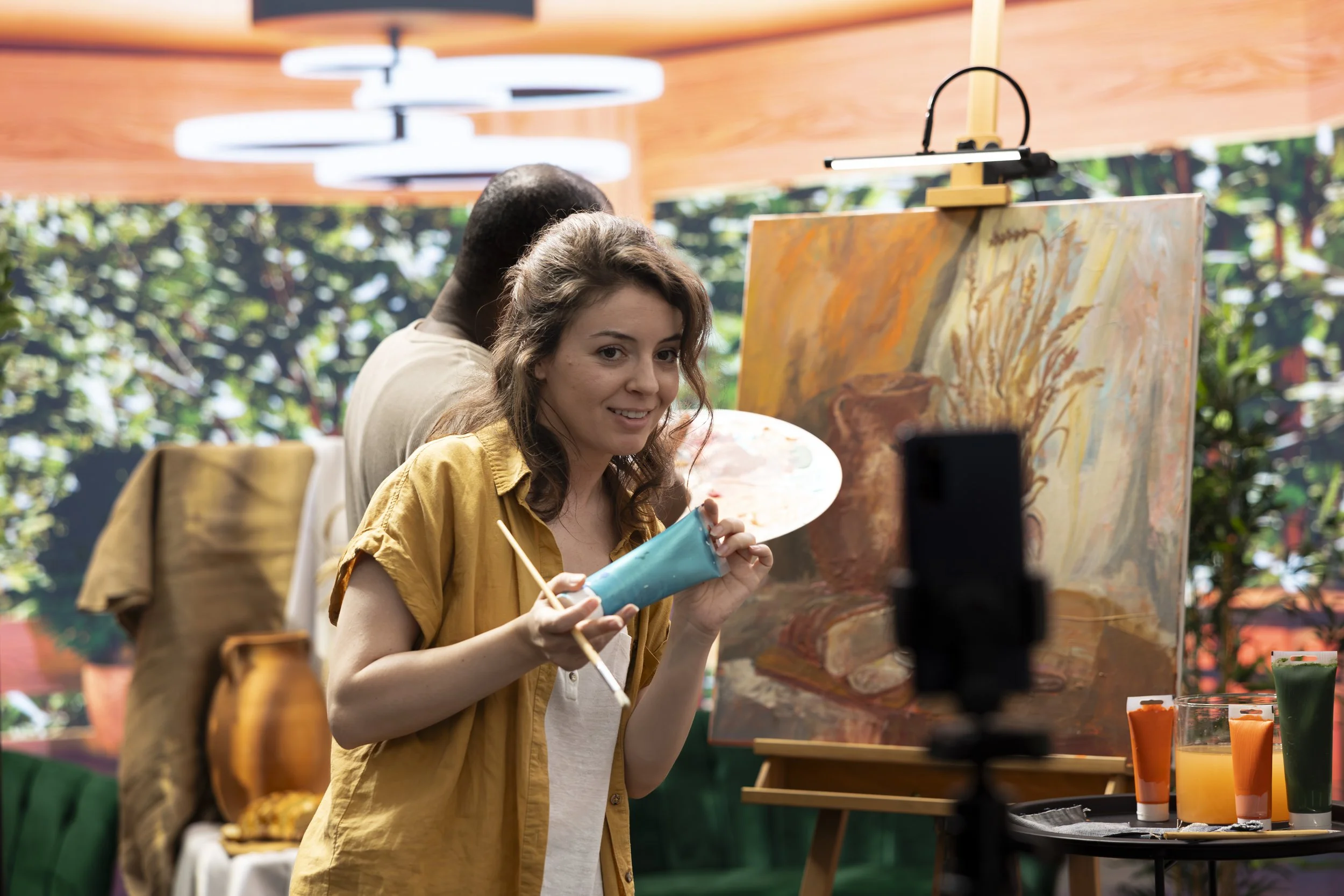 A woman holding a paintbrush and palette near an easel with an abstract painting, indoors with an orange ceiling and a camera on a tripod in front of her.
