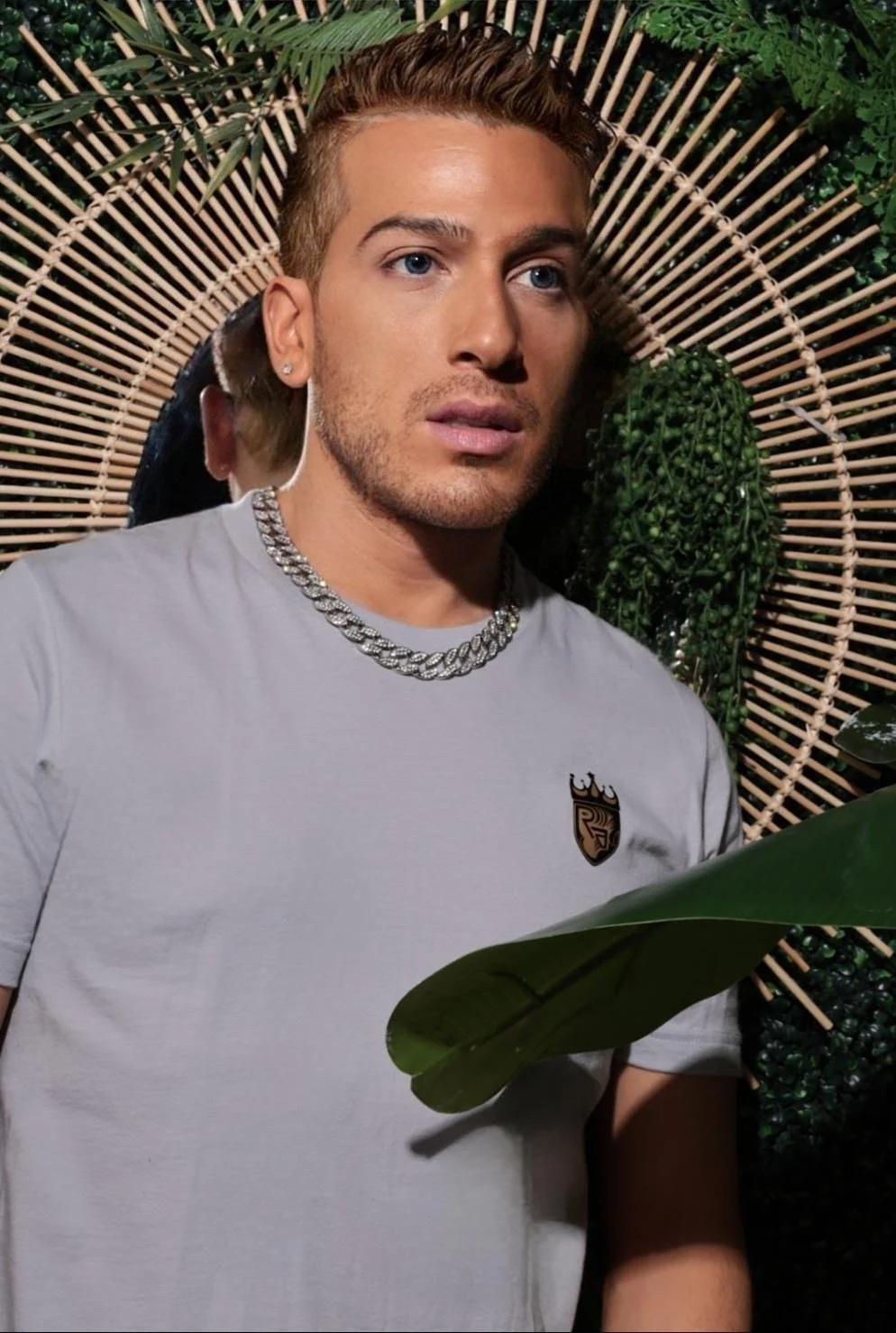 A young man with light skin, styled short hair, and a beard, wearing a white T-shirt, a thick silver chain necklace, and a small earring. He is sitting in front of a decorative background composed of green plants, a woven circular frame, and large leaves, creating a tropical ambiance.