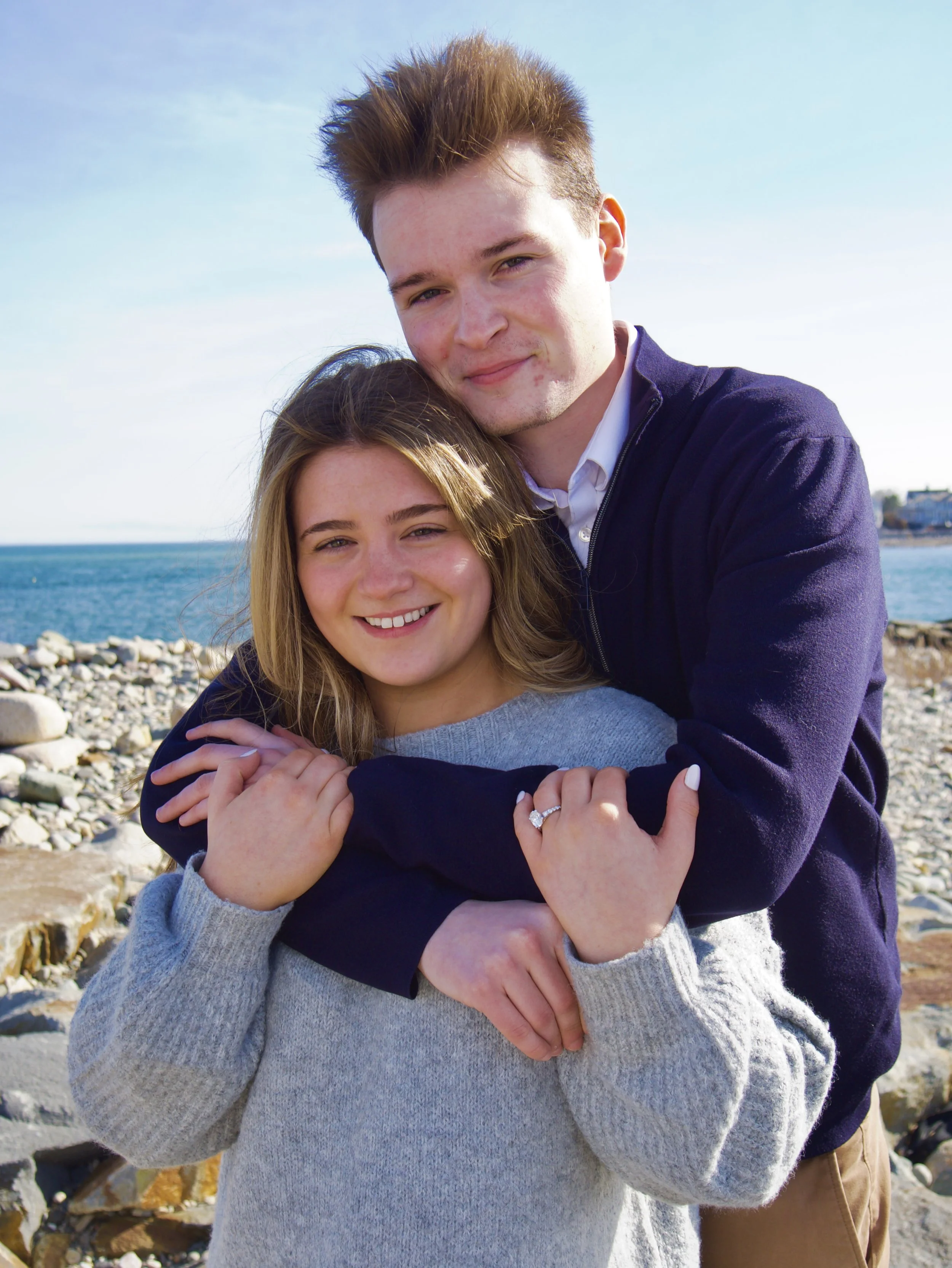 A young couple smiling and hugging on a rocky beach with the ocean and a clear blue sky in the background.