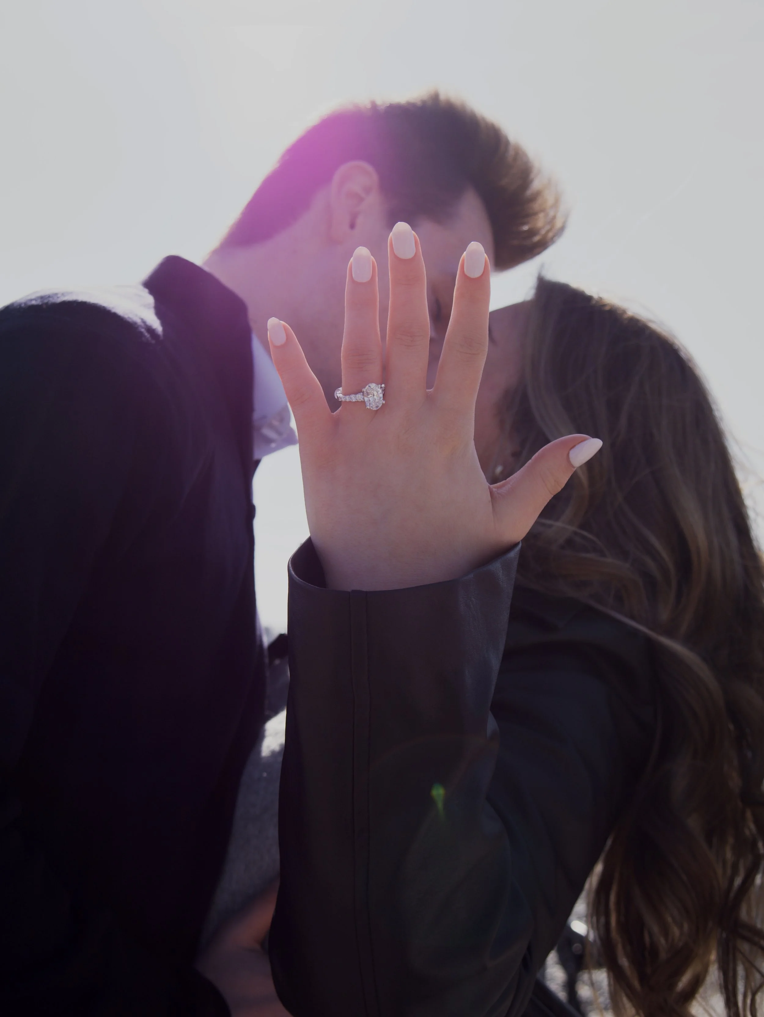 A woman showing her left hand with a diamond engagement ring, a man is kissing her forehead in the background, both are outdoors with bright sunlight.