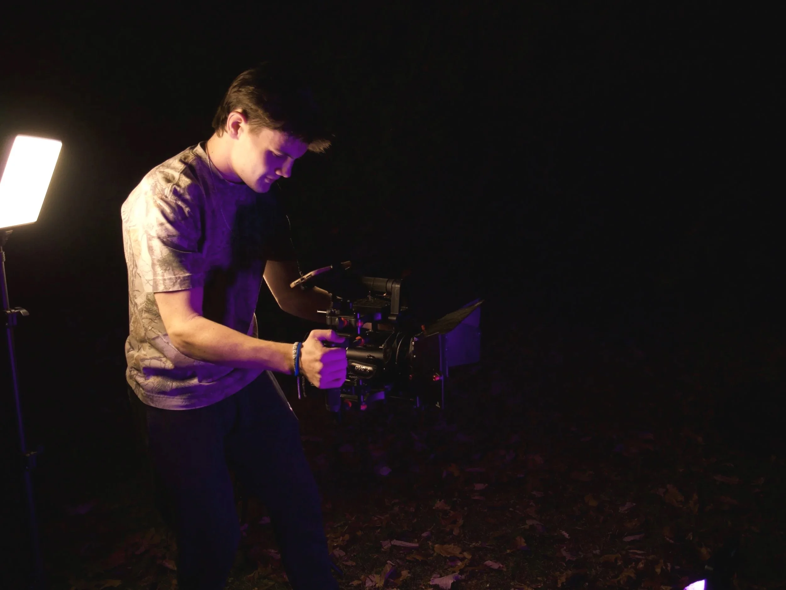 A person operating a professional camera in a dark setting with purple lighting and a bright light on the left side.