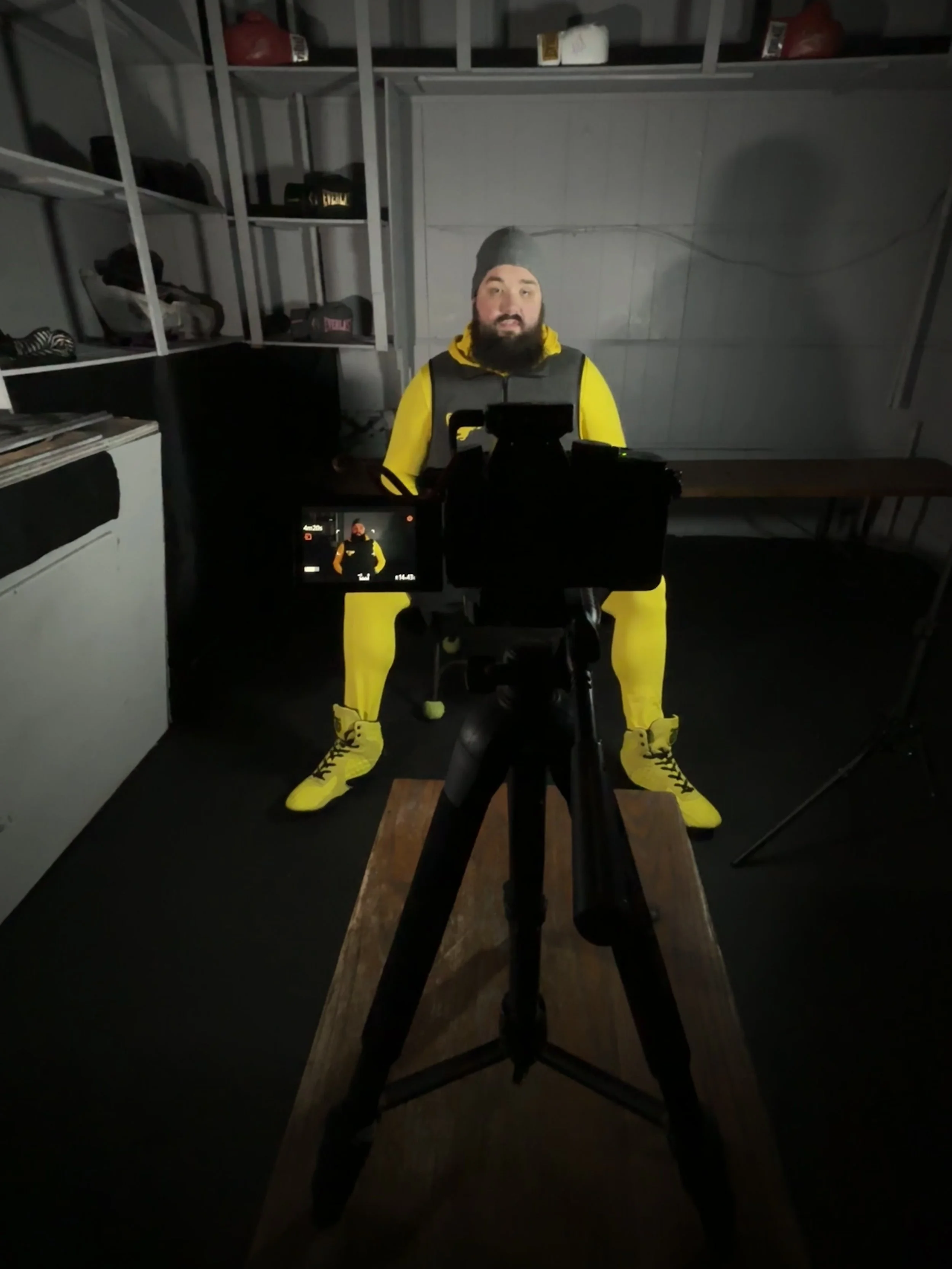 A man with a beard wearing a gray beanie, yellow jacket, and black vest sitting in front of a camera in a dimly lit room with shelves and a wooden table.