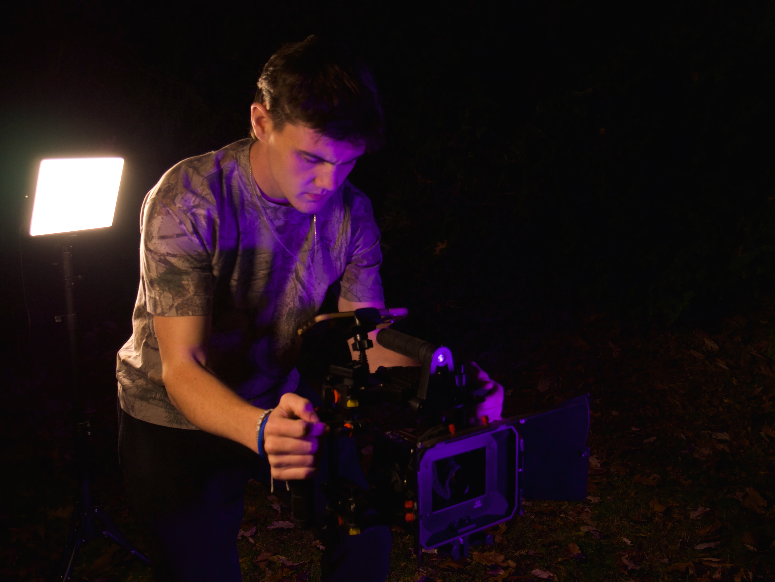 A young man working with a professional video camera outdoors at night, illuminated by purple and white lights.