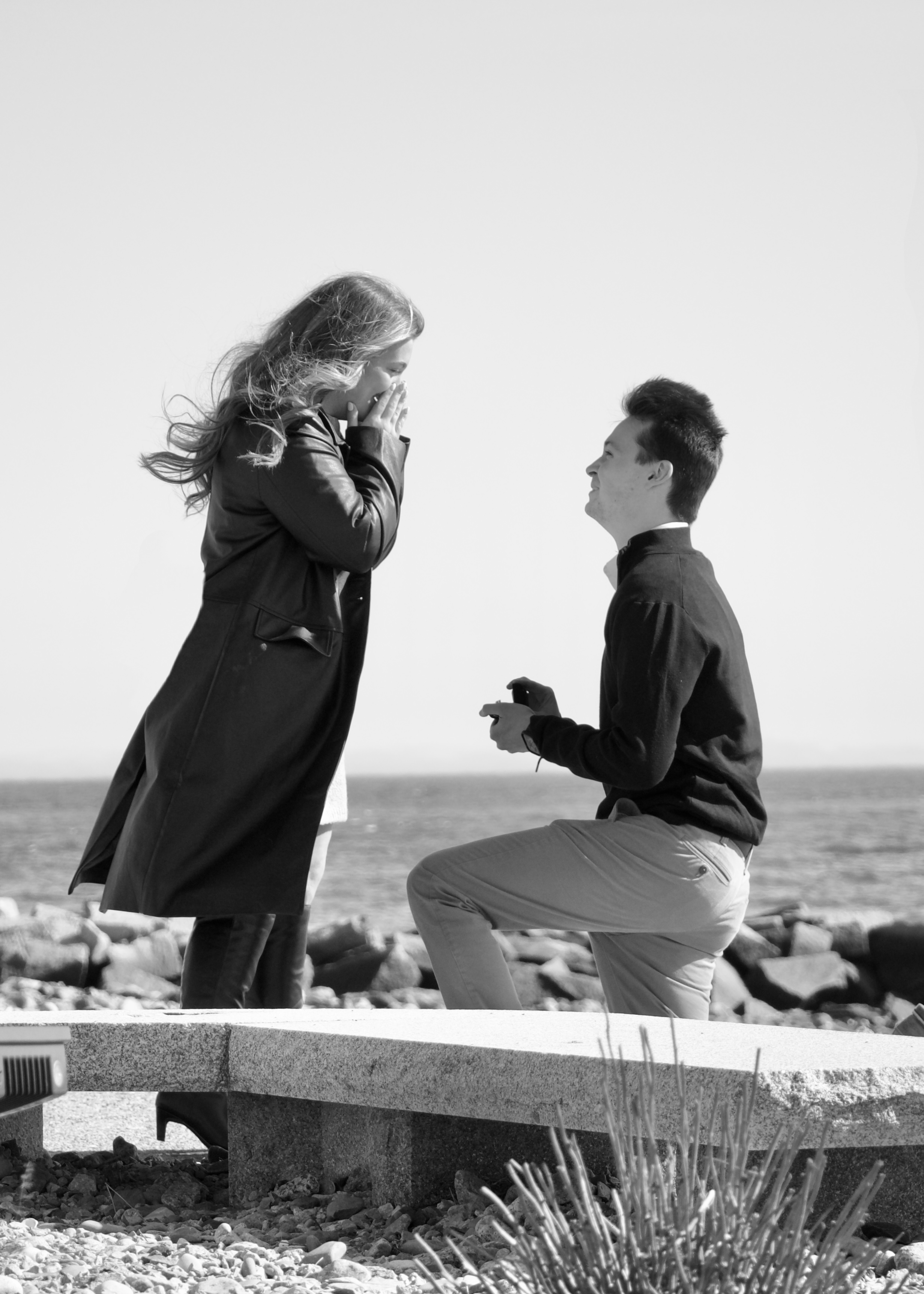 A man is proposing marriage to a woman by the beach, with both appearing emotional and happy.