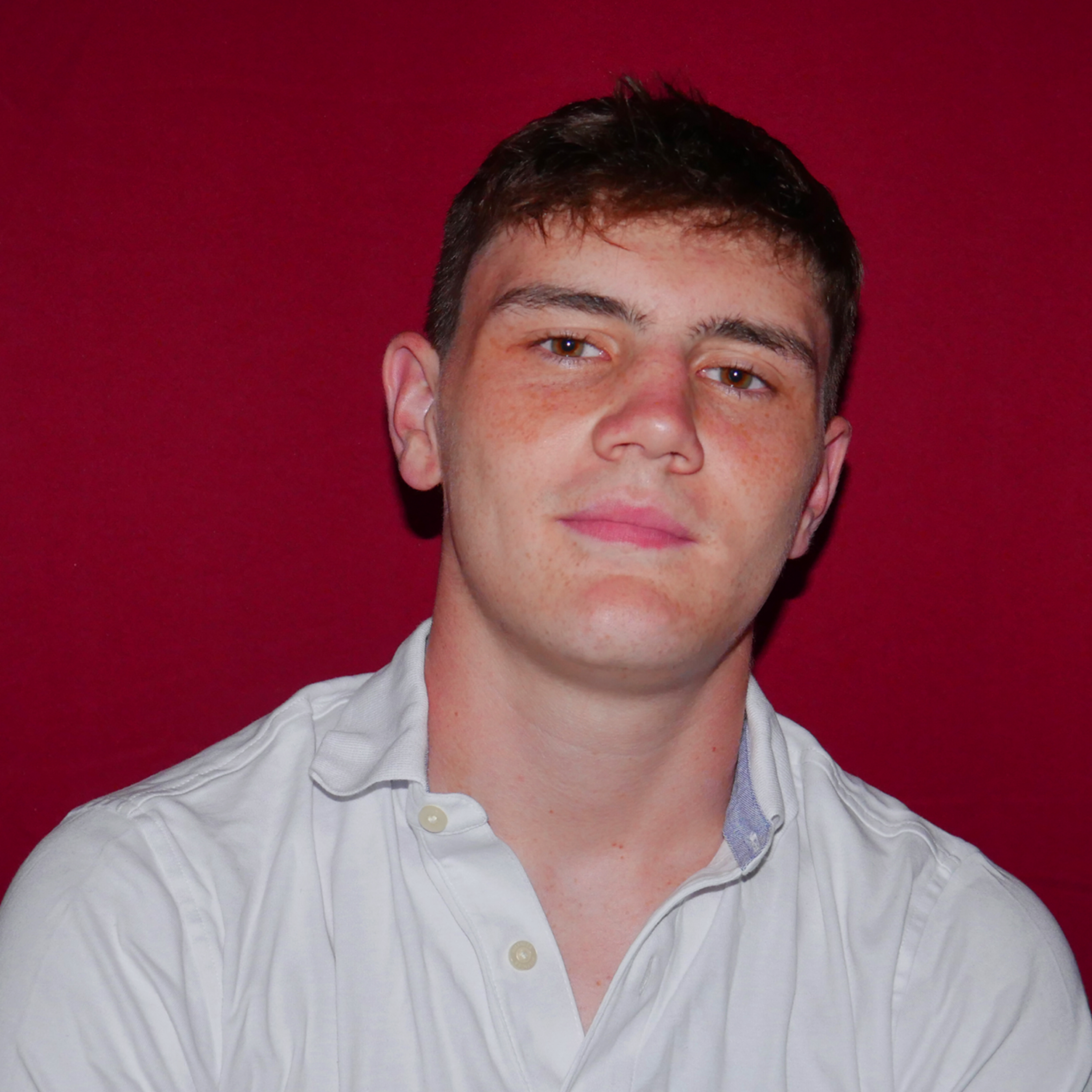 Portrait of a young man with short dark hair, wearing a white button-up shirt, against a red background.