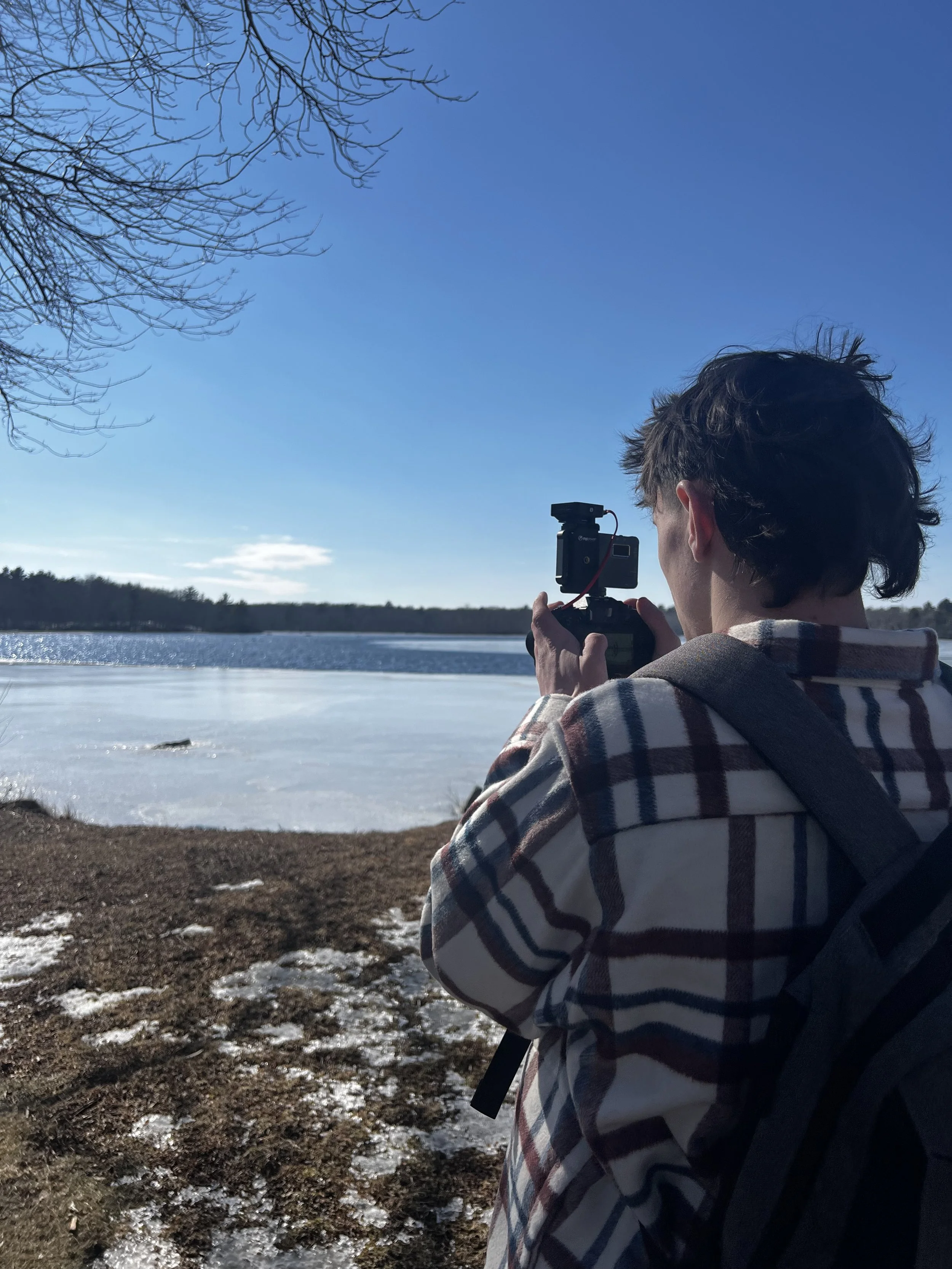 Person with a backpack and plaid jacket taking a photo of a lake with a frozen surface on a sunny day.