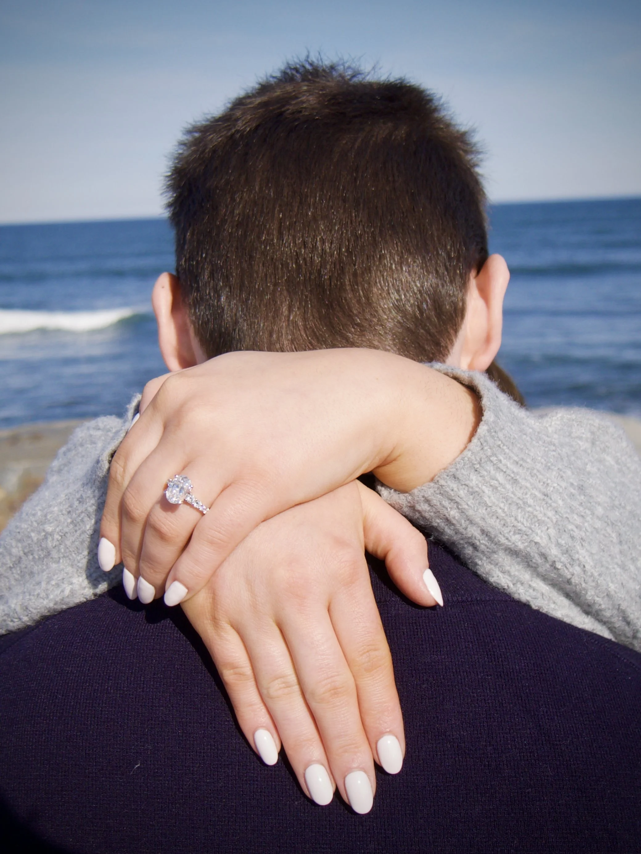 Close-up of a woman hugging a man from behind at the beach, with a focus on her hand showing an engagement ring, and waves in the background.