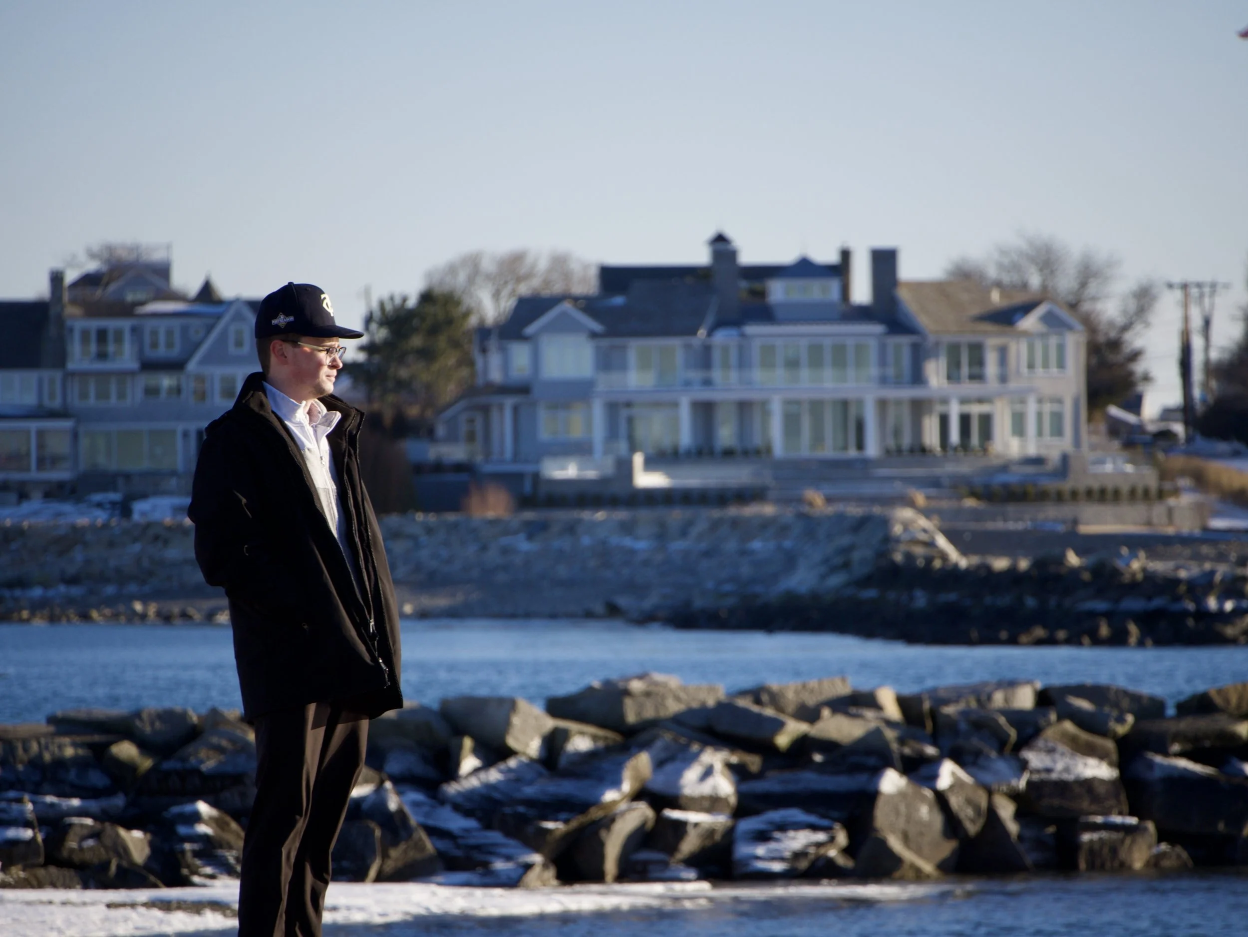 A man wearing a baseball cap, glasses, a black jacket, white shirt, and brown pants stands outdoors by the water, with large houses visible in the background on a clear, sunny day.