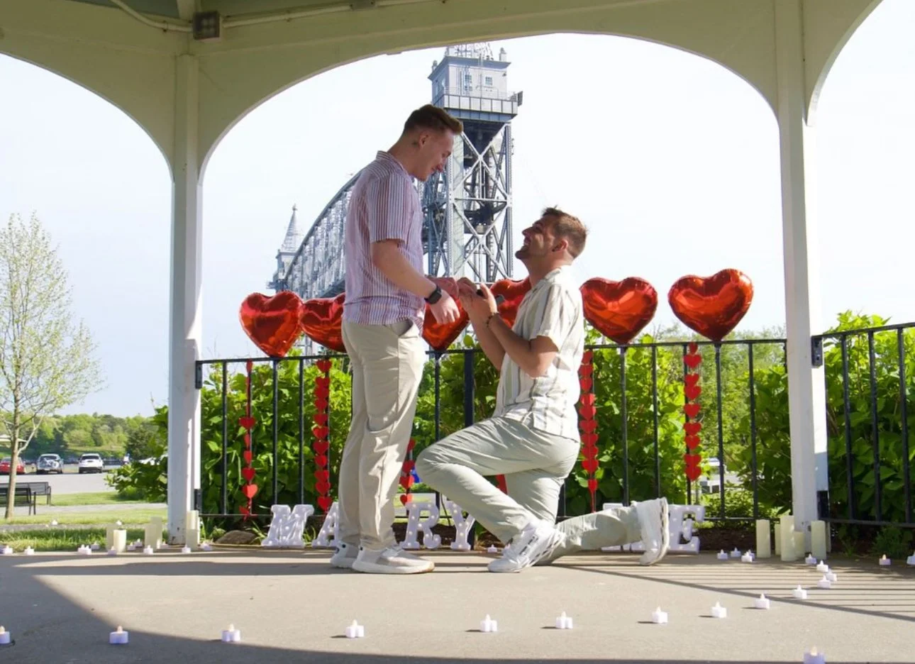 A man proposes marriage to a woman in a park pavilion decorated with red heart-shaped balloons, candles, and romantic decorations on a sunny day.
