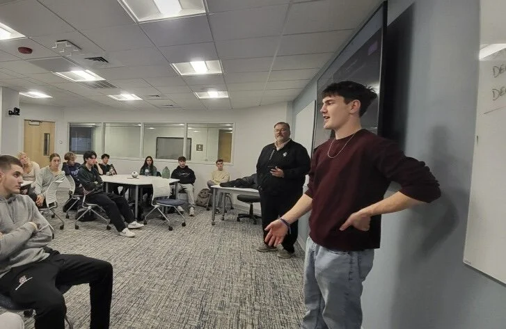 A young man is standing in front of a classroom with students seated around tables, the young man is speaking, and an older man is standing nearby.