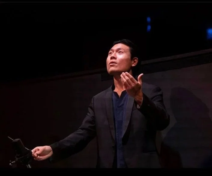A man in a dark suit gesturing with his right hand while speaking or conducting in front of a dark background, possibly at a music festival in Besançon, France.