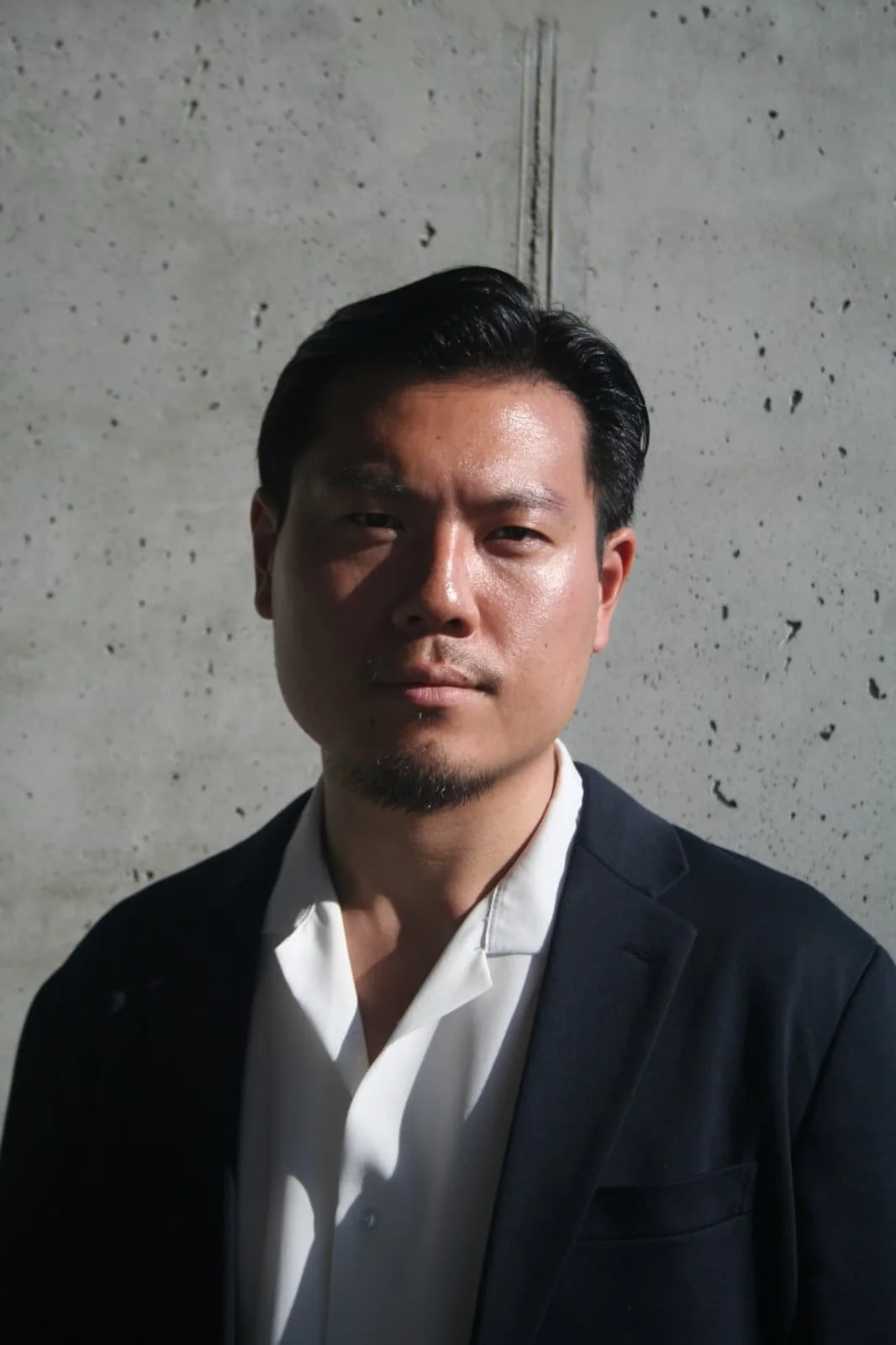 A man with dark hair and a goatee wearing a black blazer and white shirt standing against a concrete wall.