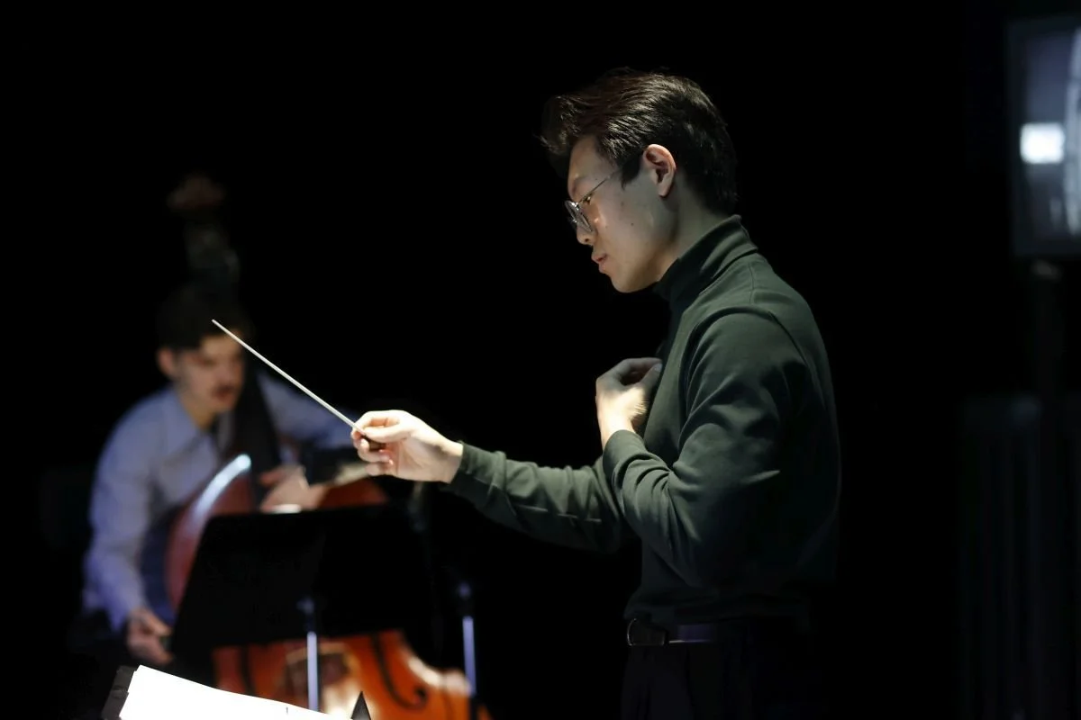A man wearing glasses and a dark green shirt conducts an orchestra with a baton, with a blurred musician playing the cello in the background.