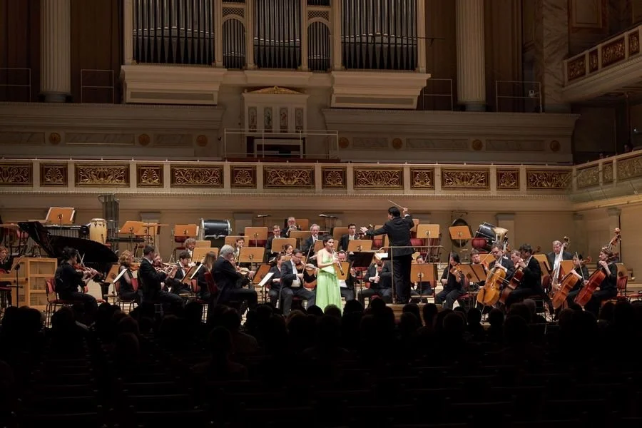 An orchestra performing on stage in a concert hall, with a female soloist in a green dress and a conductor in front of them.