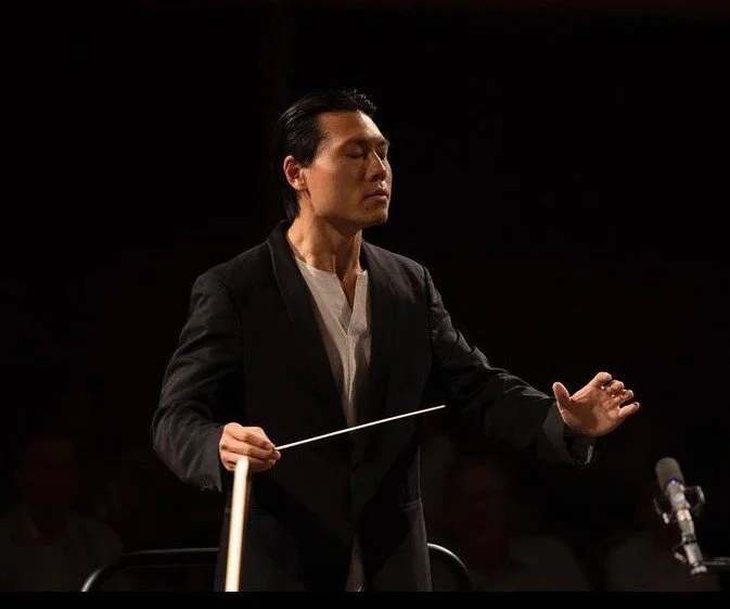 Conductor leading an orchestra, holding a baton, with eyes closed and raised hand.