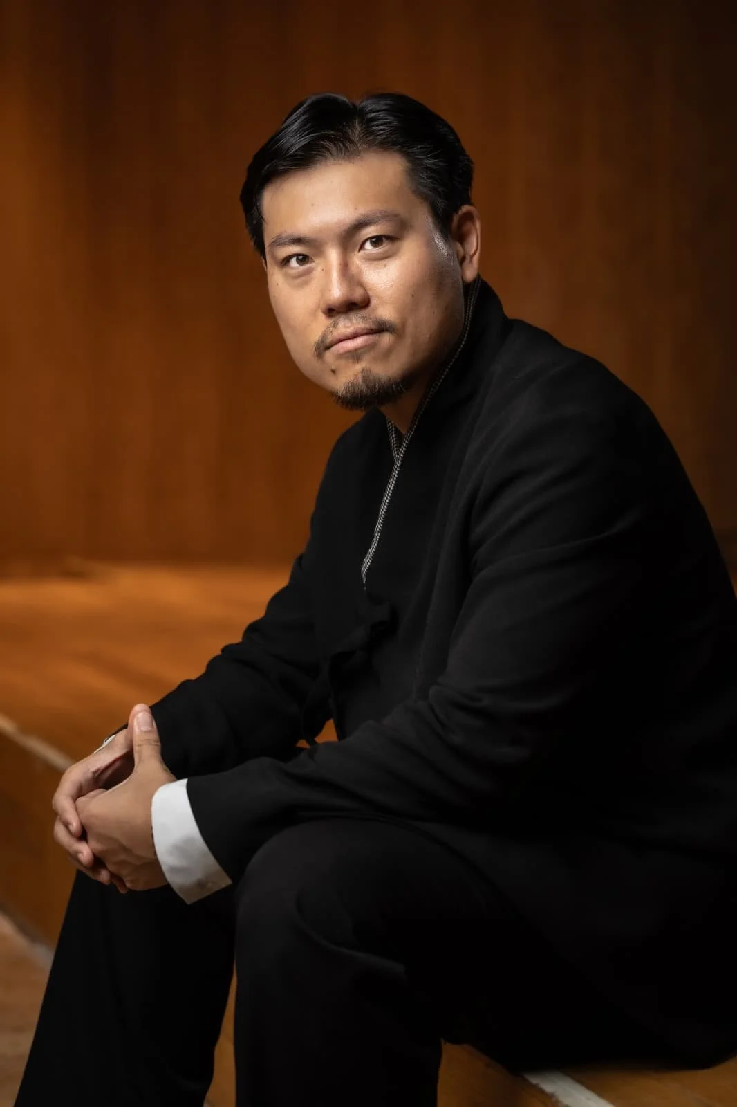 Portrait of a man with black hair and a goatee, wearing a black jacket, sitting on a wooden bench with a warm brown background.