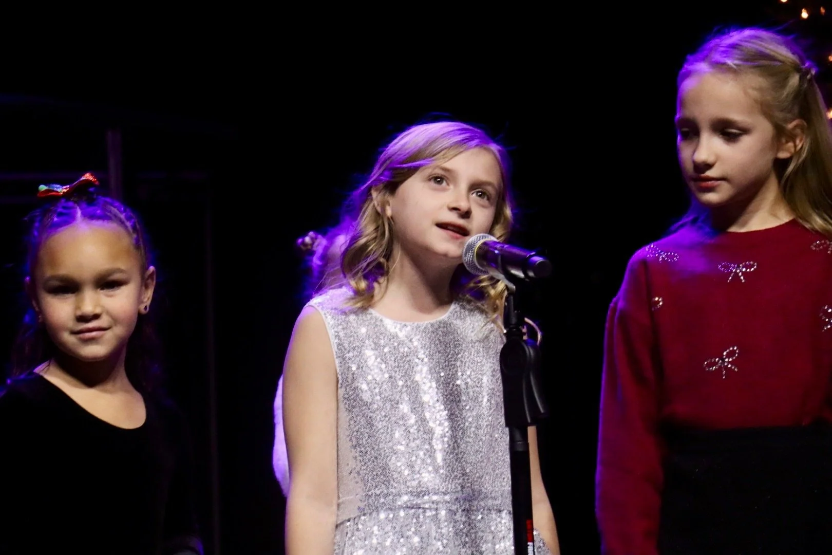 Three young girls standing on stage, one speaking into a microphone, lit with stage lighting against a dark background.