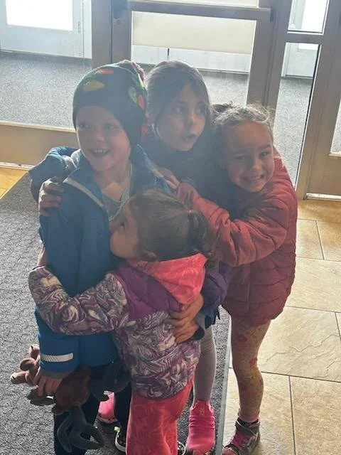 Four children hugging and smiling near a glass door, dressed in warm clothes.