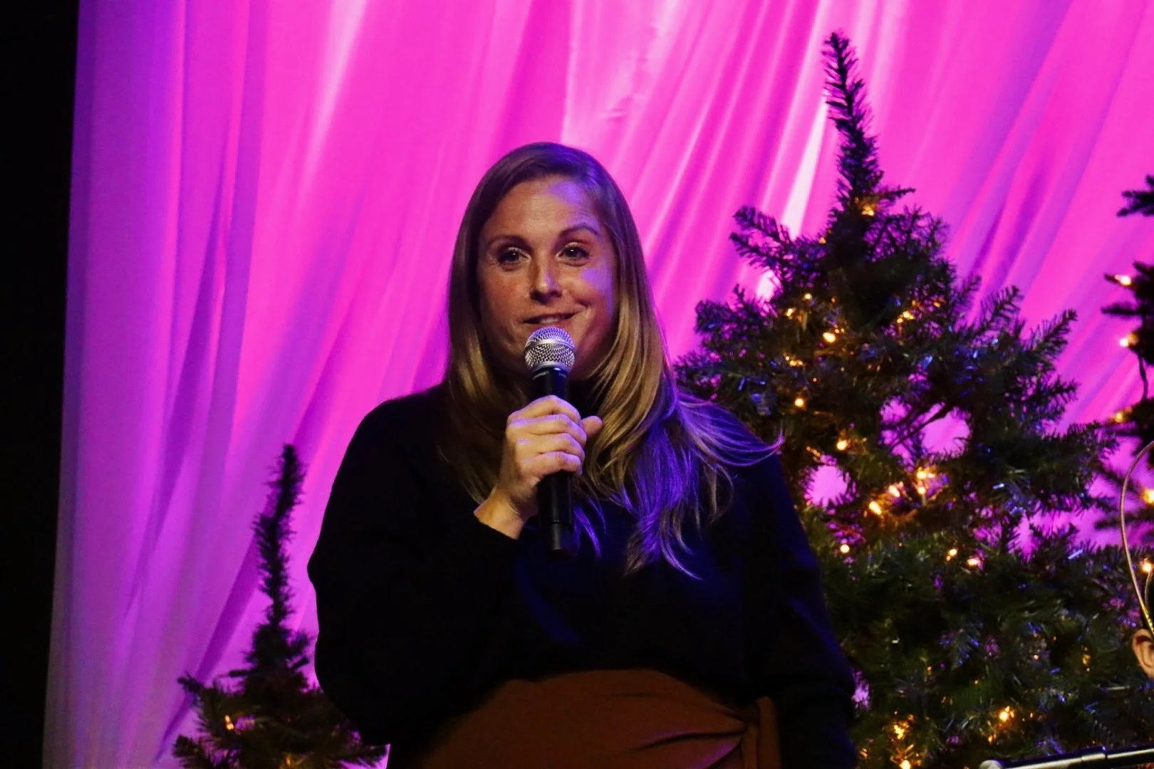 Woman with long blonde hair holding a microphone, standing in front of pink and purple draped background with decorated Christmas trees.