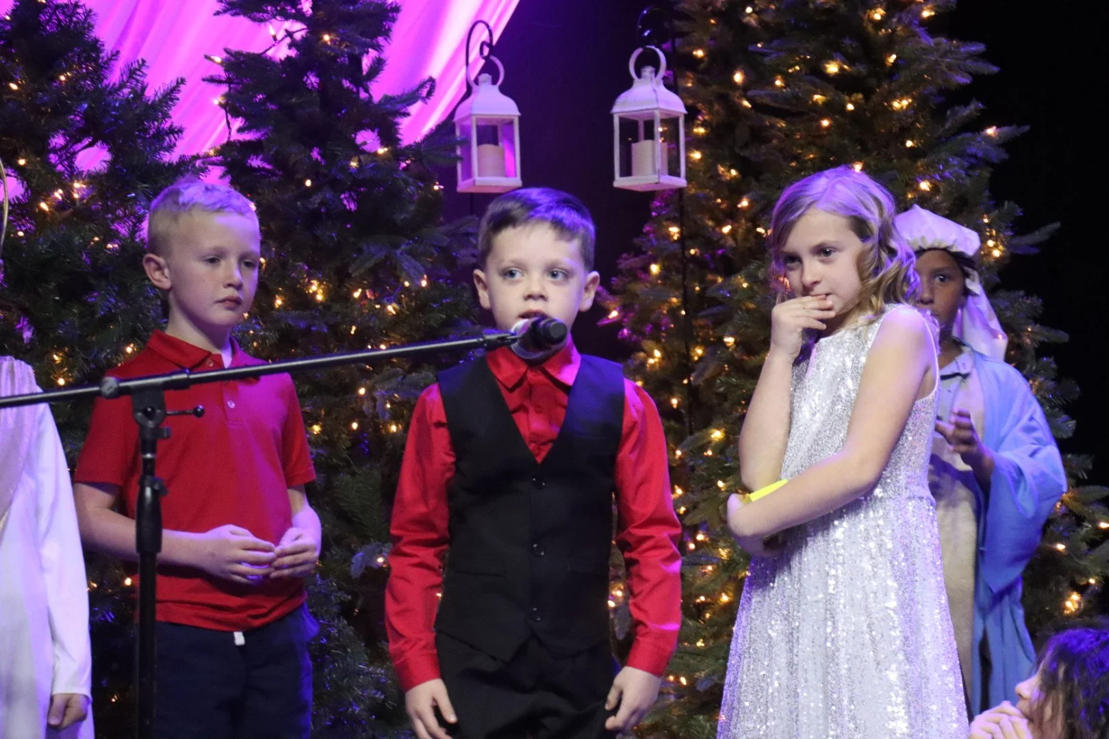 Children performing on stage during a Christmas play, with decorated Christmas trees and lanterns in the background.