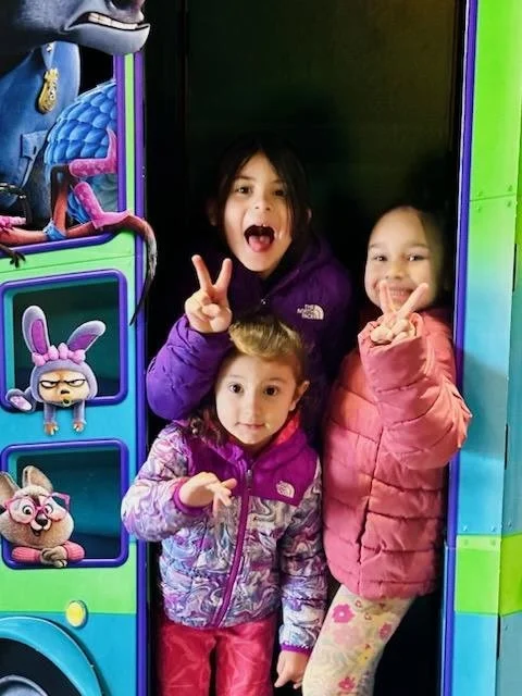Three young girls smiling and making peace signs while standing inside a colorful arcade or play area.