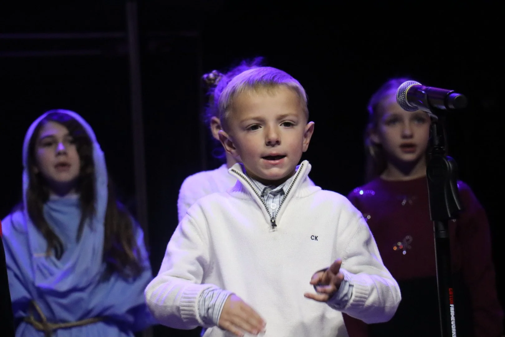 Three children perform on stage with a microphone, the boy in the center wearing a white sweater, and two girls, one wearing a hooded outfit and the other in a red top with decorations.
