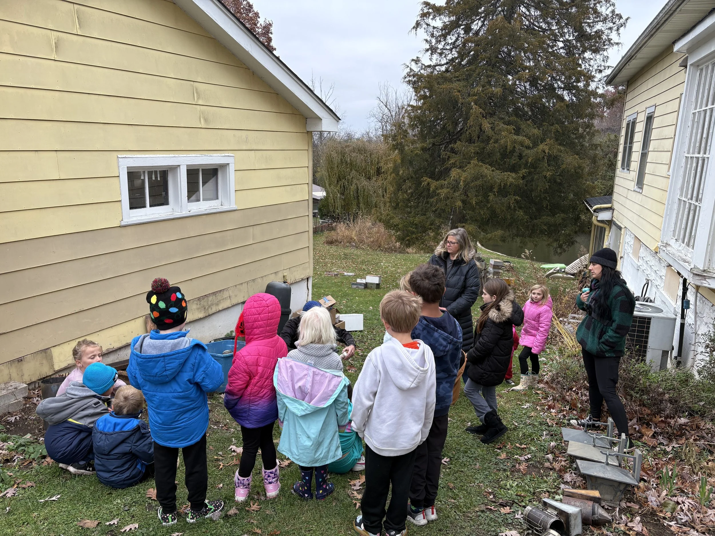 A group of children and two women gathered outside near yellow and white houses, listening to a person who appears to be giving a demonstration or talk, in the yard with fallen leaves and some garden items.