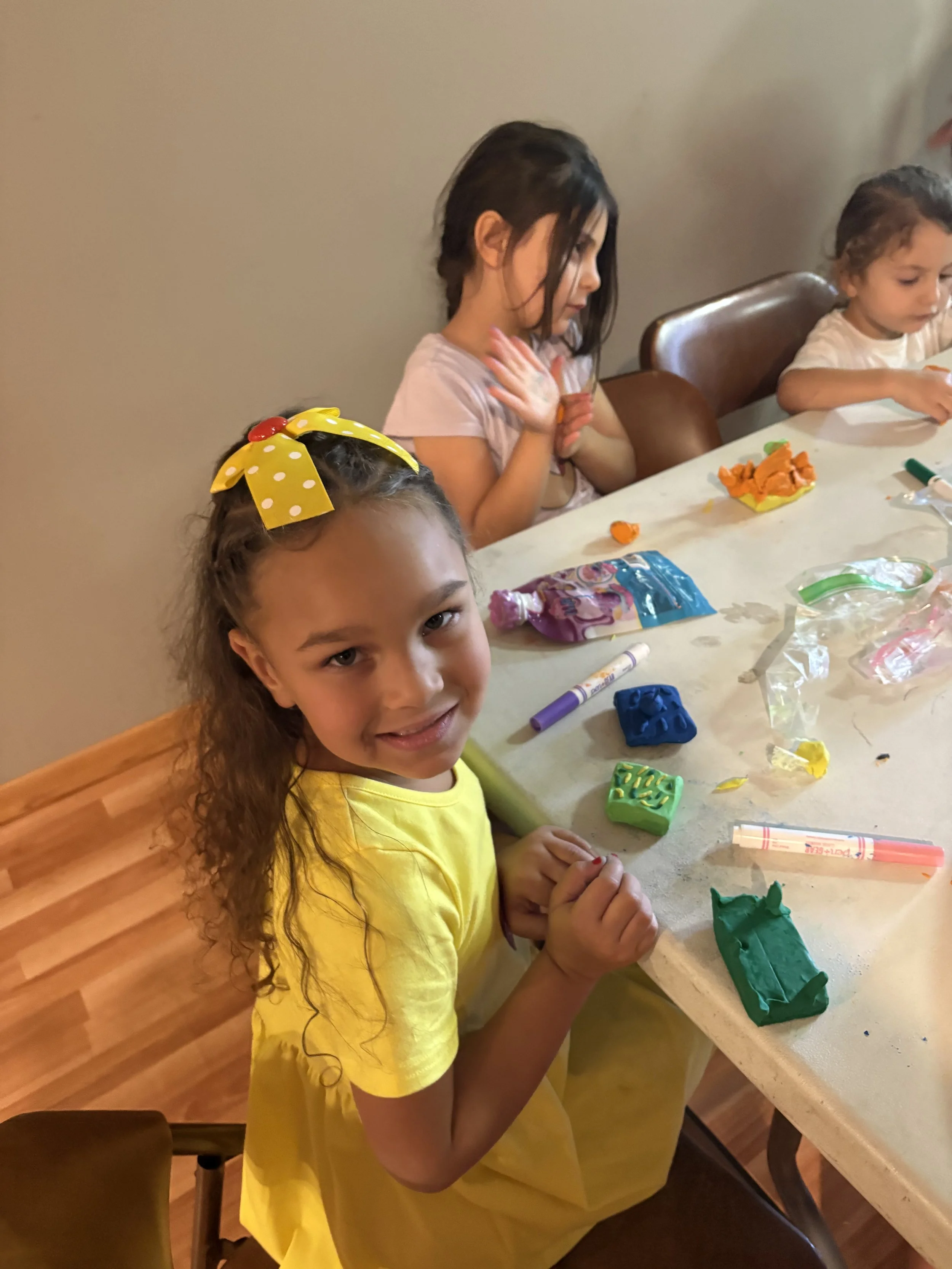 A young girl with curly hair and a yellow bow sitting at a table with modeling clay and art supplies, smiling at the camera; two other children are in the background, also engaging with art materials.