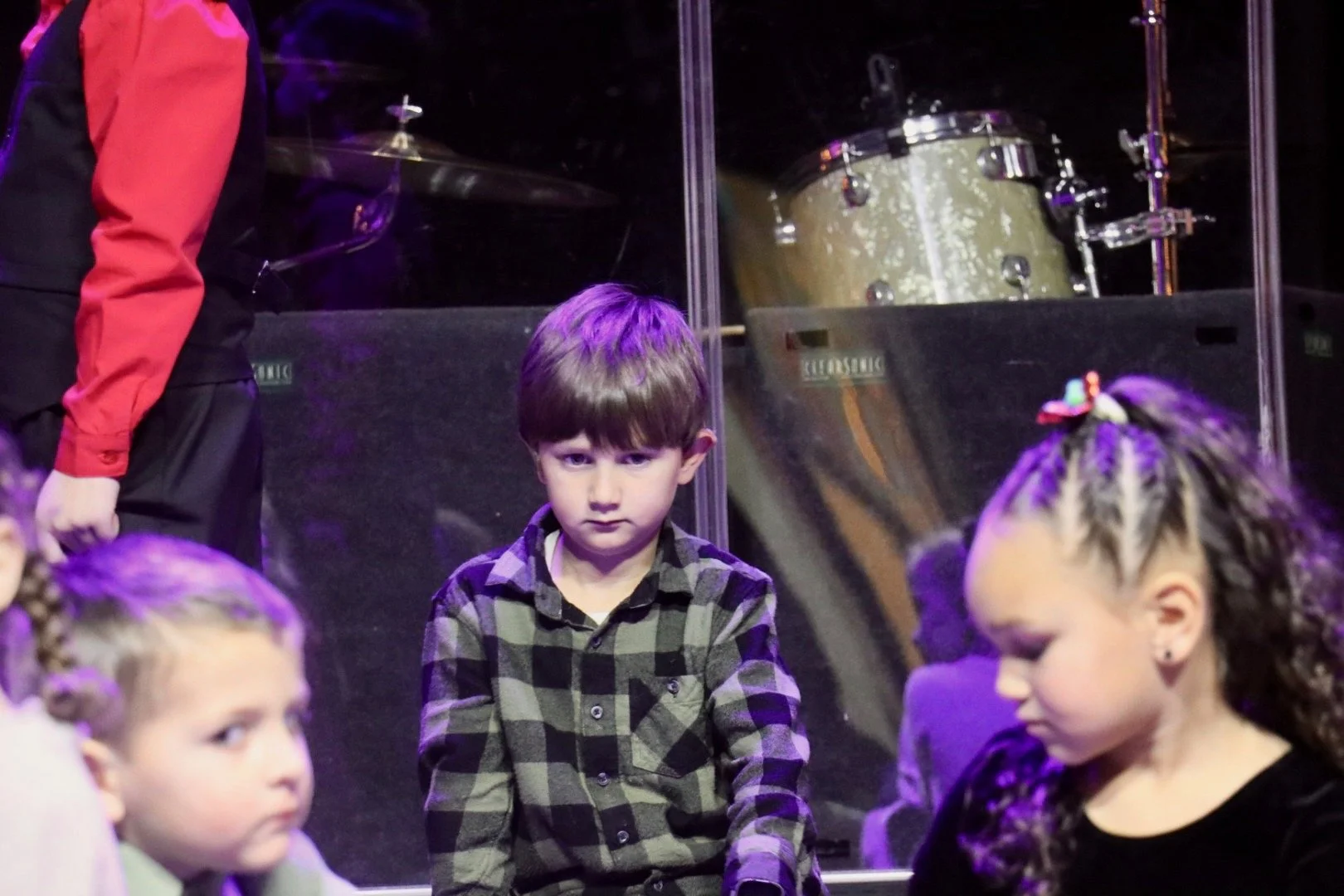 Young boy in plaid shirt looking at camera in front of a glass partition, with children and drum set visible behind it.