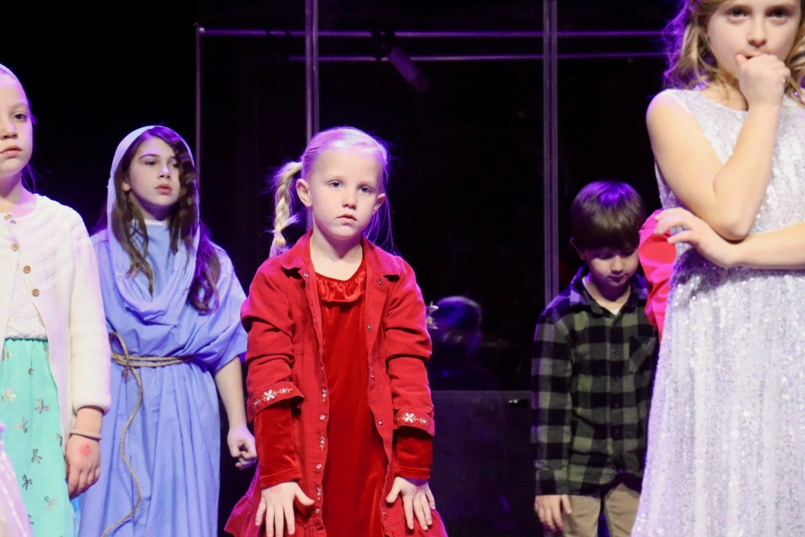Children standing on stage during a theatrical performance, with a girl in a red dress in the center and other children around her.