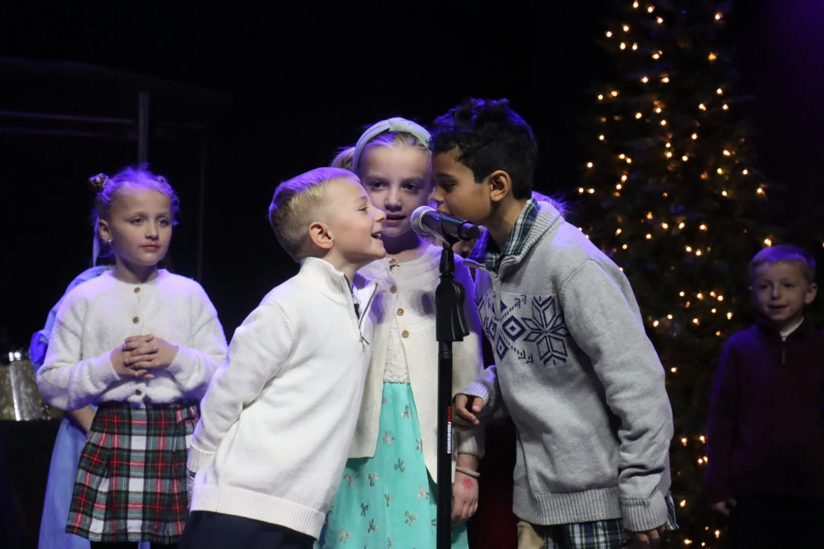 Children performing Christmas carol on stage, with Christmas tree in background.
