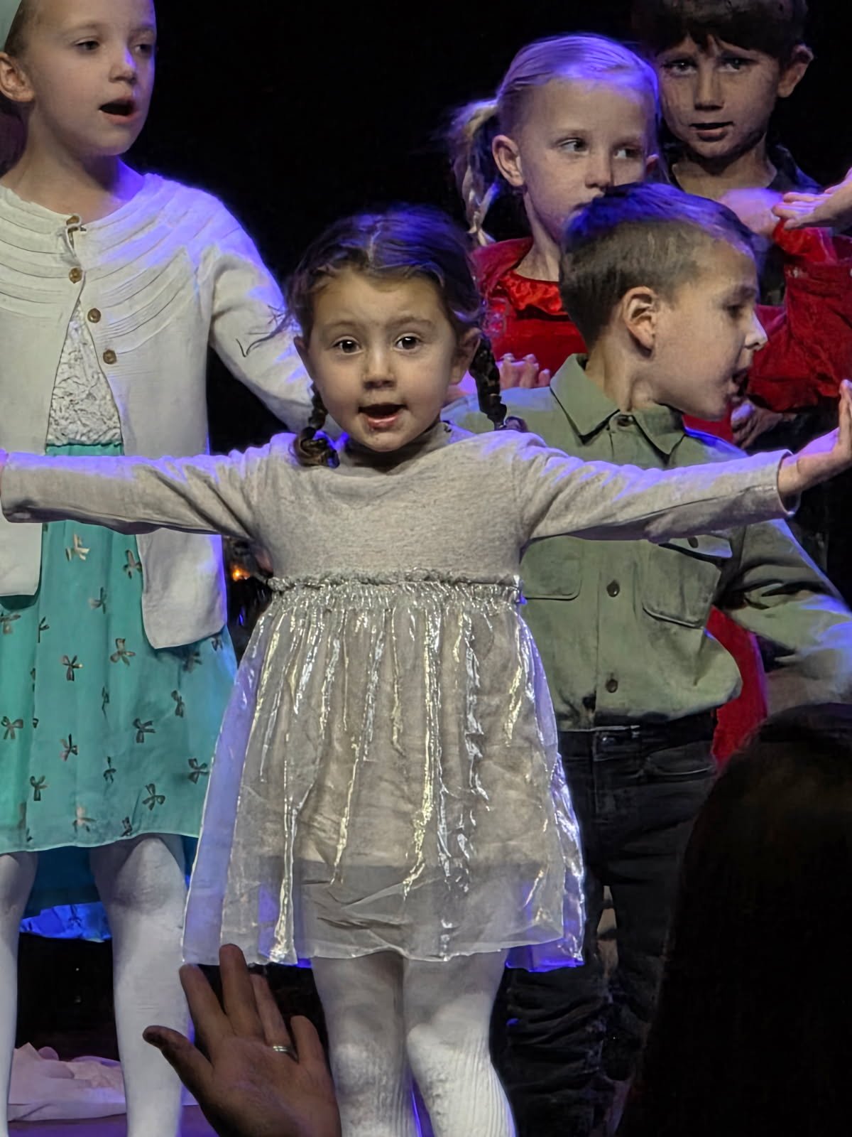 Children on stage performing, with girl in front wearing a silver dress and white tights, stretching her arms out, surrounded by other children in colorful costumes.