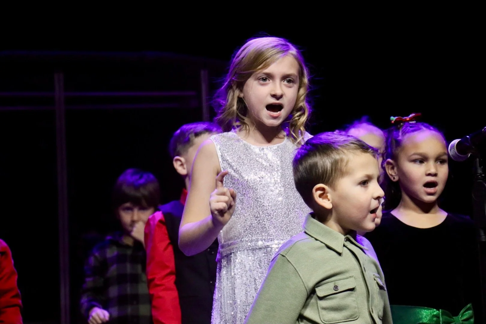 Children performing on stage with one girl singing into a microphone, wearing a shiny silver dress, and others standing behind her.