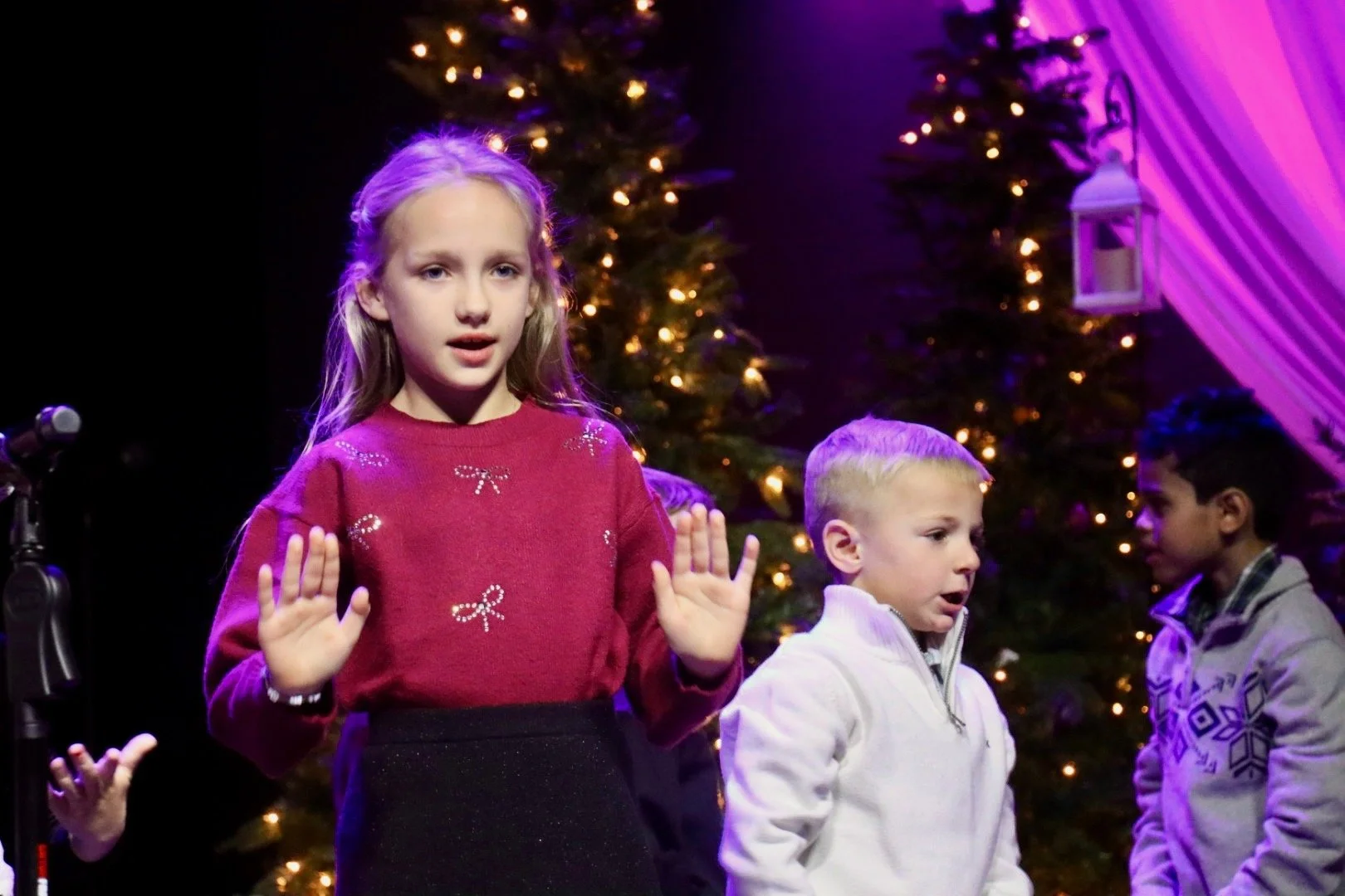 Three children standing in front of decorated Christmas trees with fairy lights, during a Christmas event. The girl in the red sweater is in the foreground with hands raised, and two boys are alongside her.