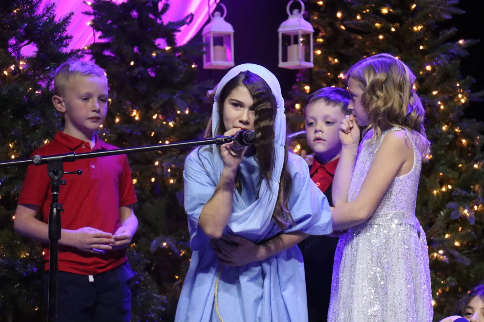Children performing a nativity scene on stage with Christmas trees and lights in the background.