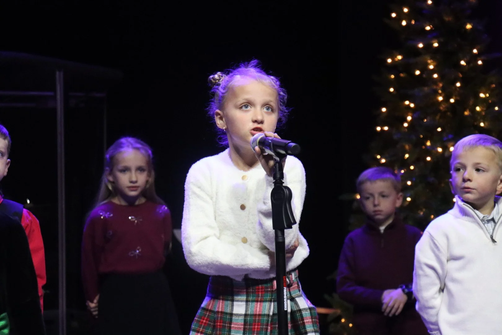 Young girl in a white sweater and plaid skirt speaking into a microphone during a Christmas event, with other children and a decorated Christmas tree in the background.