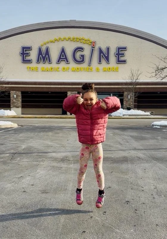 A young girl in a pink jacket and floral leggings jumping in front of an Emagine movie theater building, giving a thumbs-up and smiling.